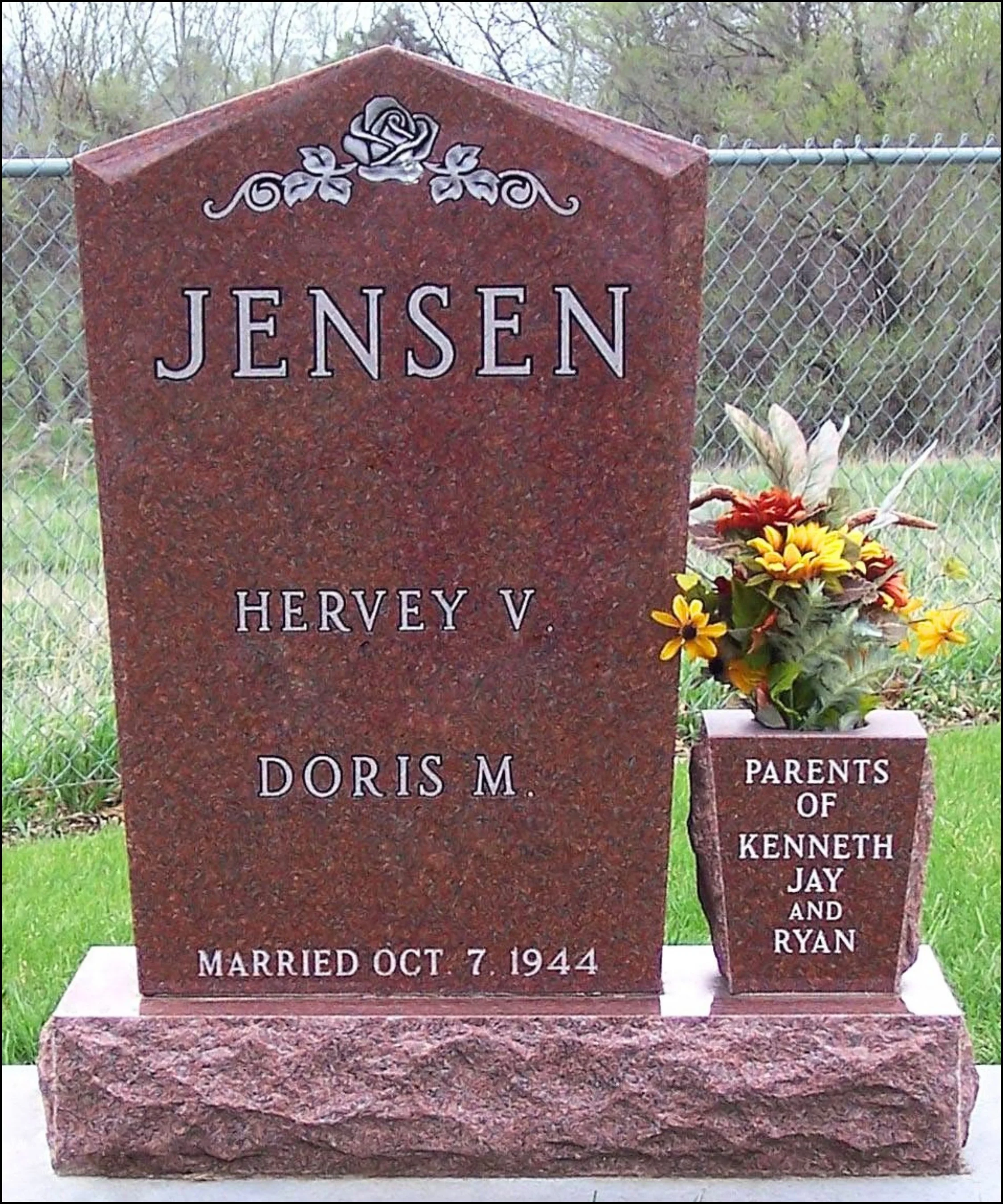 A red granite headstone with engraved and painted white text, commemorating Doris M. Jensen, her wedding date, and her children, with a small floral arrangement next to it on a grassy area with trees and a chain-link fence in the background.