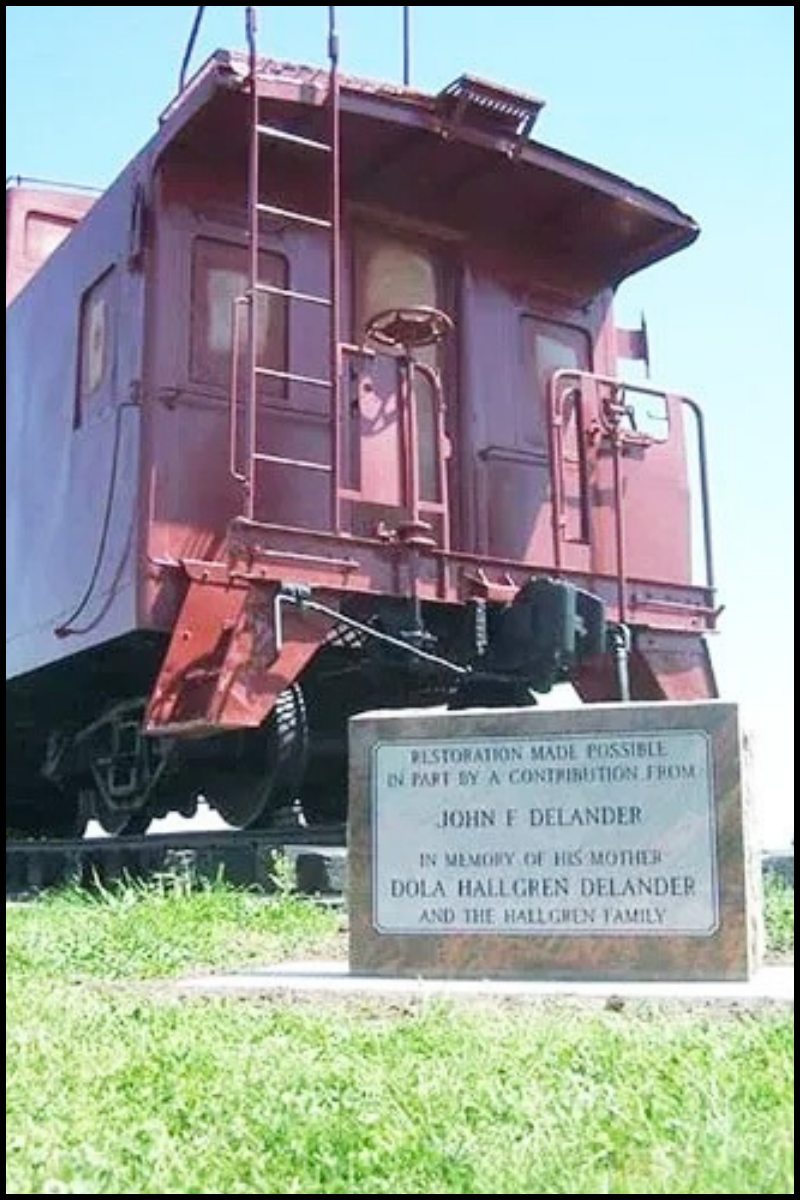 A vintage train locomotive on display with a memorial plaque in front of it. The plaque reads: "Restoration made possible in part by a contribution from John E Delander in memory of his mother Dola Hallgren Delander and the Hallgren family."
