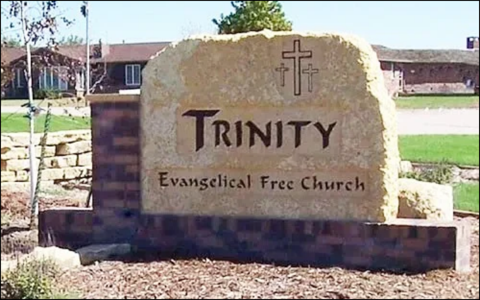 A stone sign with the word 'TRINITY' and 'Evangelical Free Church' engraved on it, with three crosses depicted above the word.