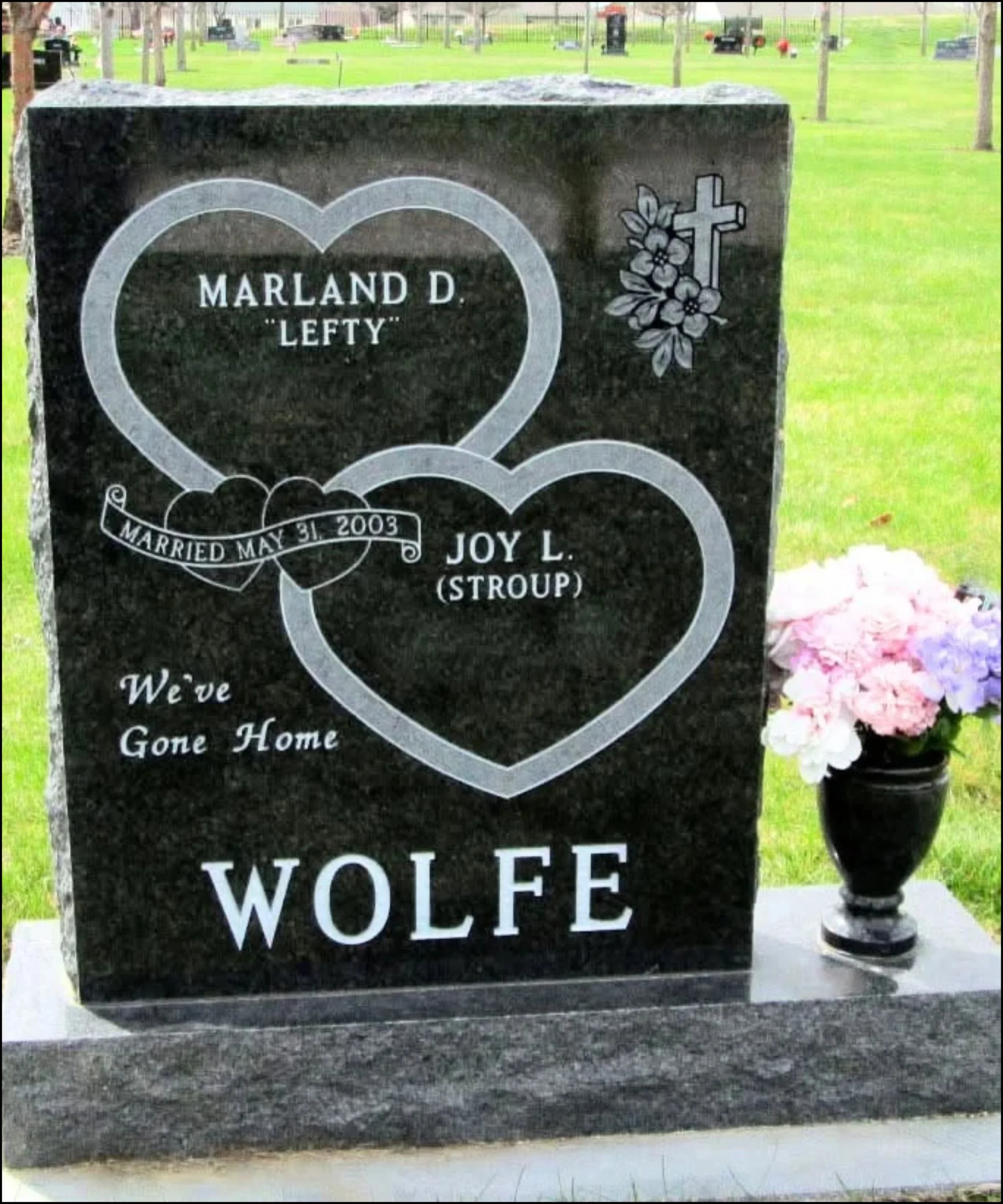 Close-up of a black granite headstone with white engraved text reading "Marland D. 'Lefty'" and a large heart outline. The cemetery has green grass, trees, and other gravestones in the background, with a small bouquet of pink and white flowers in a black vase beside the headstone.
