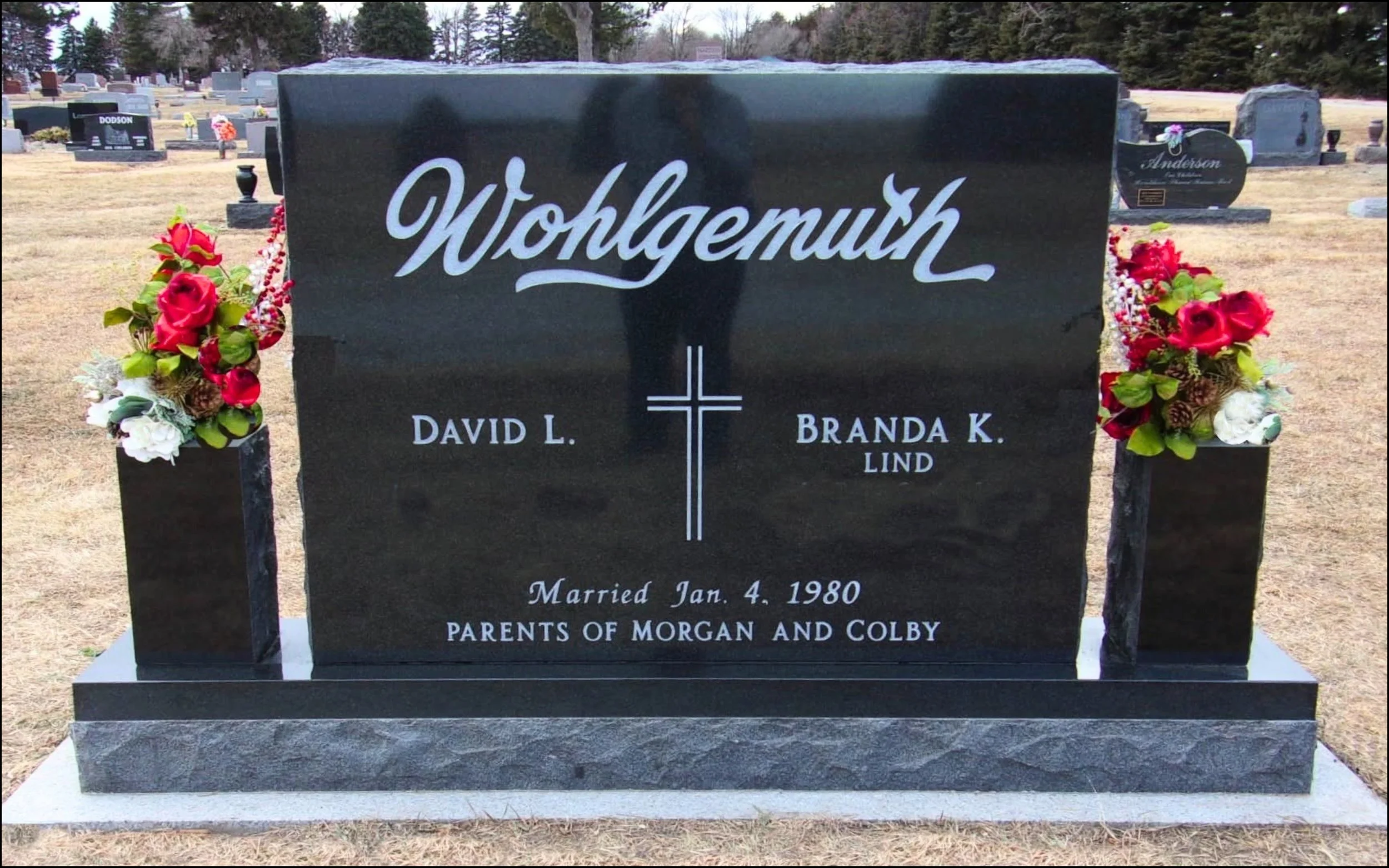 Black granite headstone with white engraving and floral arrangements on either side, in a cemetery. The headstone bears the name 'Wolfgemuth' and includes details of marriage, children, and death.