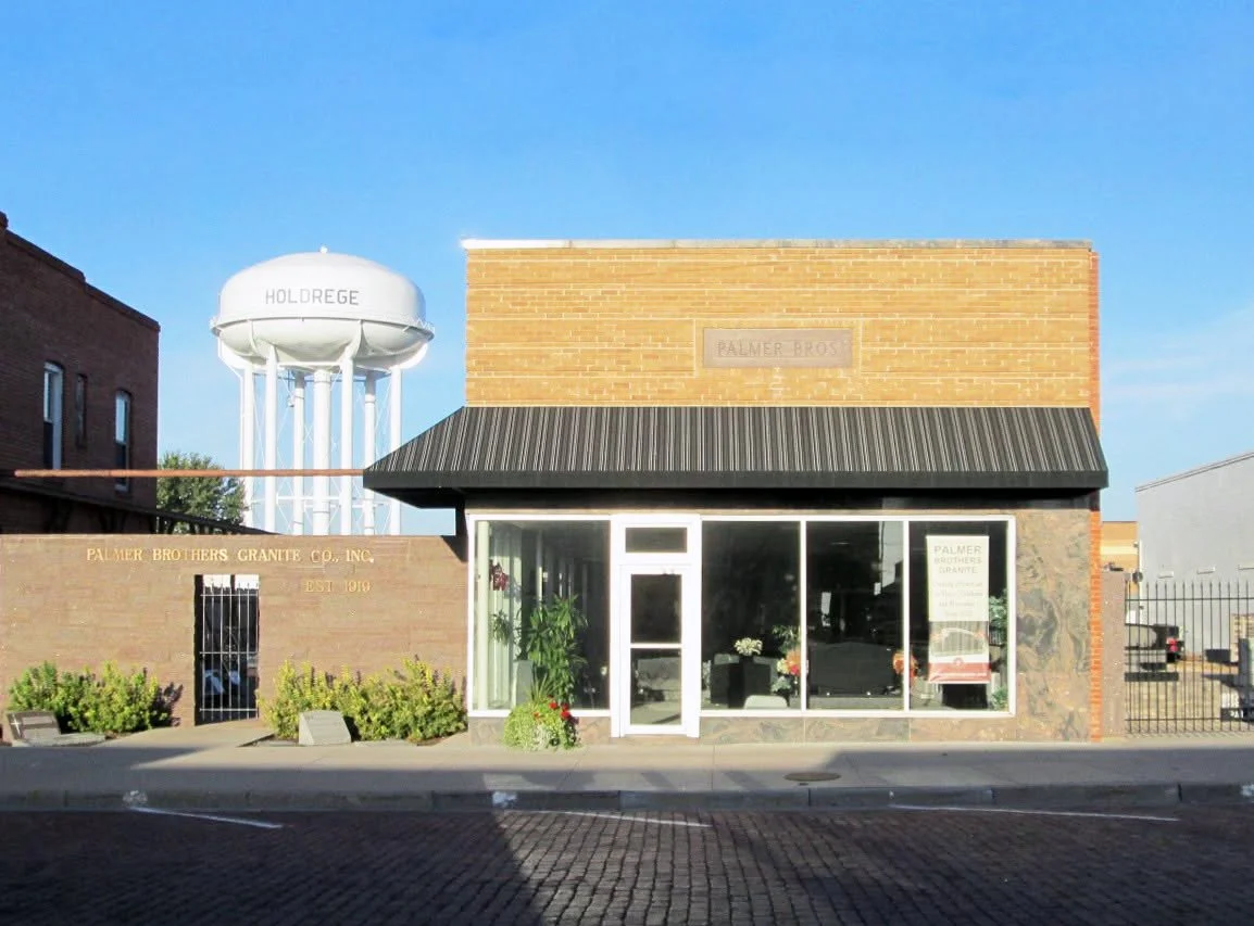 Storefront with glass windows, plants inside, brick facade, and a black awning in front. A water tower labeled 'Holdrege' can be seen in the background.
