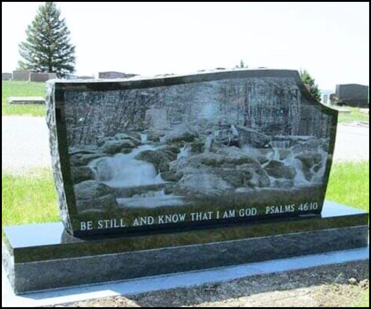 A polished black granite headstone with a carved river scene featuring rocks and trees. An inscribed biblical verse at the bottom reads, "Be still, and know that I am God. Psalms 46:10."