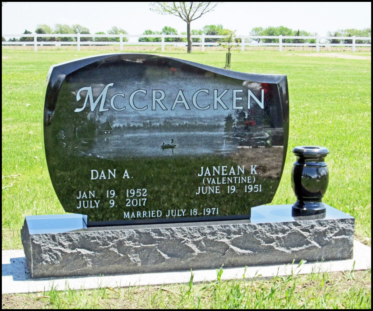 Black granite headstone with the name McCracken engraved, along with names Dan A. and Janean K. Valentine, their birth and death dates, and their wedding date, positioned on a grassy area with a tree and fence in the background.
