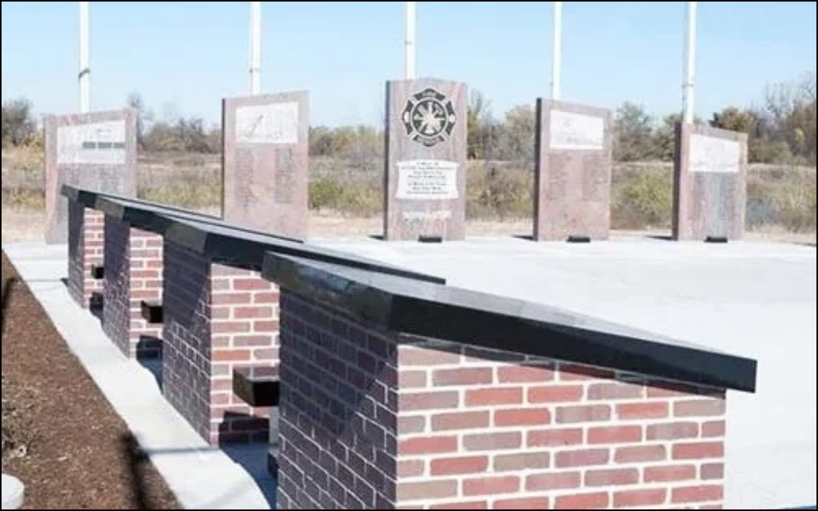 Memorial plaza with six brick pillars with plaques and a metal rail, set in an outdoor area with a clear sky and sparse trees in the background.