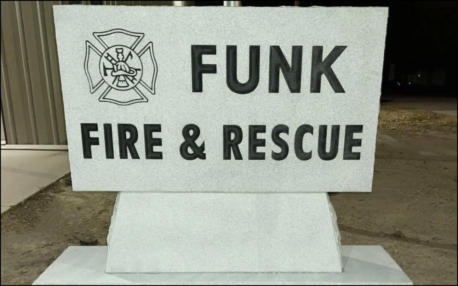 A white sign with black text that reads "FUNK FIRE & RESCUE" and features a fire department emblem, situated on a sidewalk outside at night.