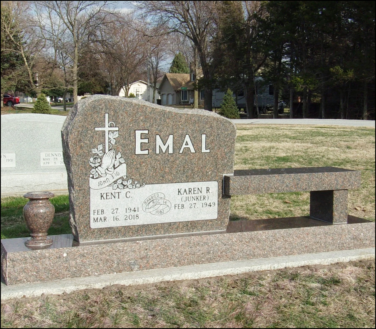 Gravestone for Kent C. Emal and Karen R. (Junker) Emal in a cemetery, with a bench and a small vase in front of it, trees and houses in the background.