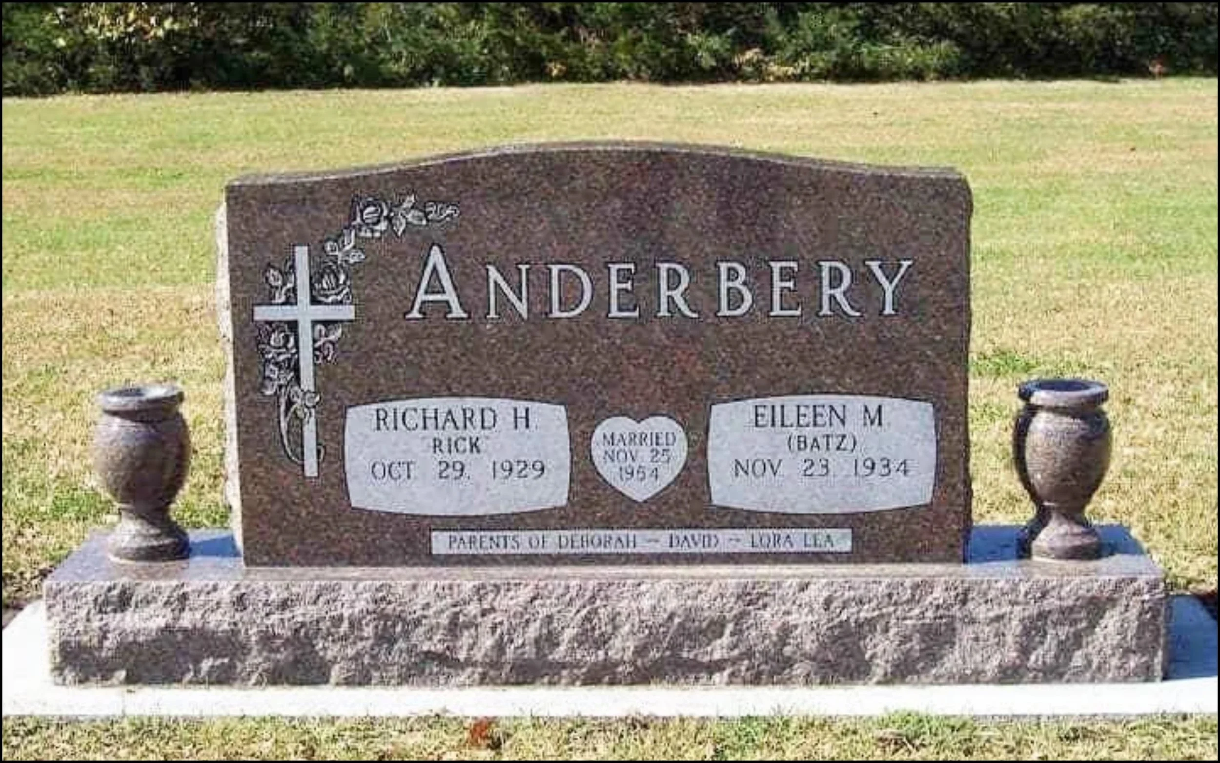 Gravestone with the family name Anderbery, featuring a cross and floral design on the top left corner. Names of Richard H. and Eileen M. Anderbery along with their birth and death dates are engraved on the stone. Smaller sign indicates the parents of Deborah, Dads, Lora, and Lea, with surrounding grass and a background of trees.