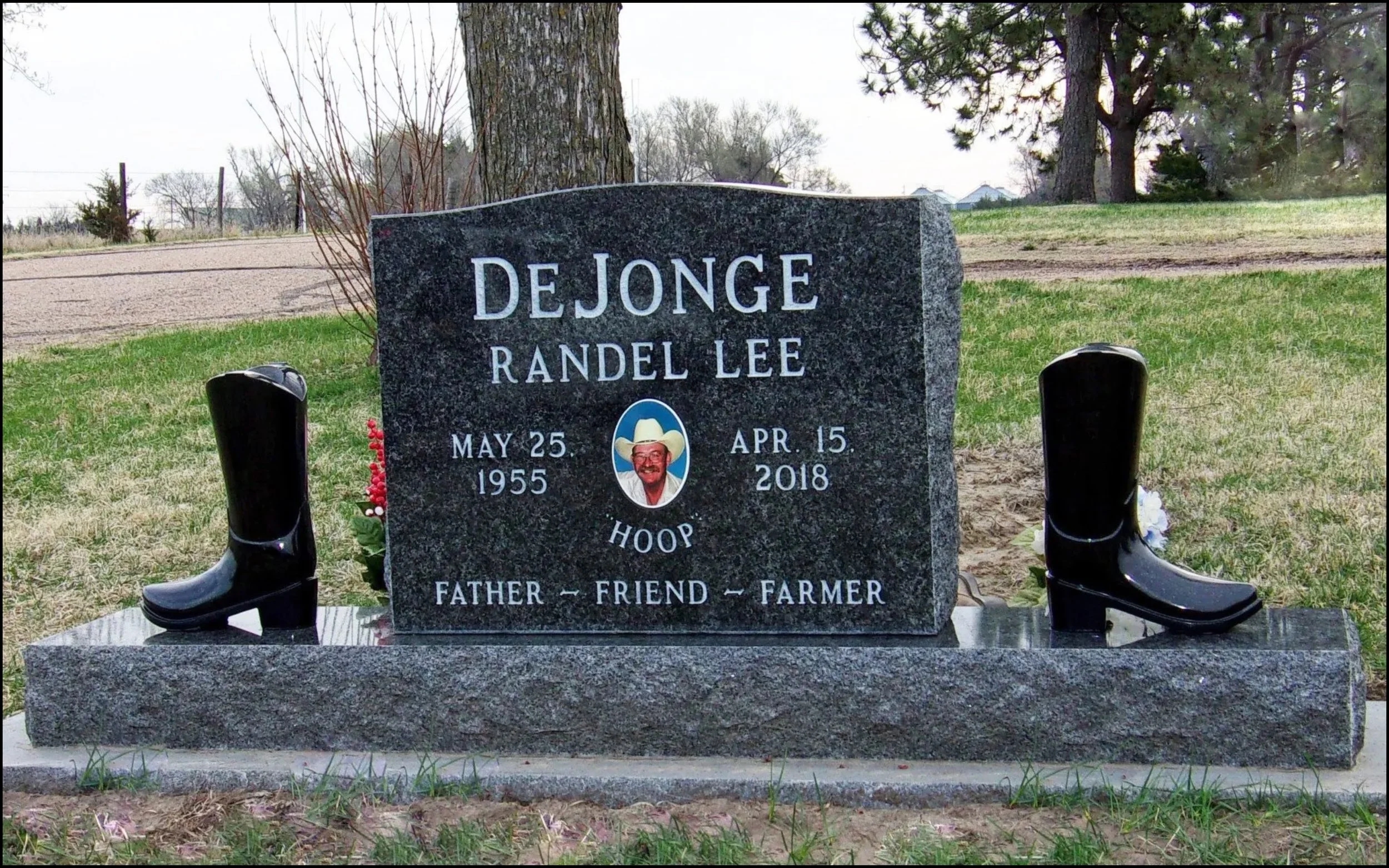 Gravestone for Randell Lee DeJonge with a photo of a man in a cowboy hat, date of birth May 25, 1955, and date of death April 15, 2018, with boots on both sides and small flowers at the base.
