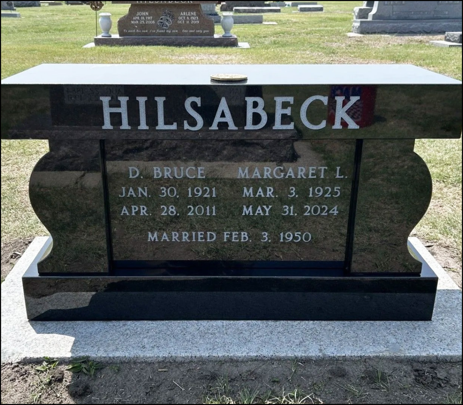 Black granite headstone for Hilsa Beck, inscribed with name, birth and death dates, and marriage date, located in a cemetery with other headstones in the background.