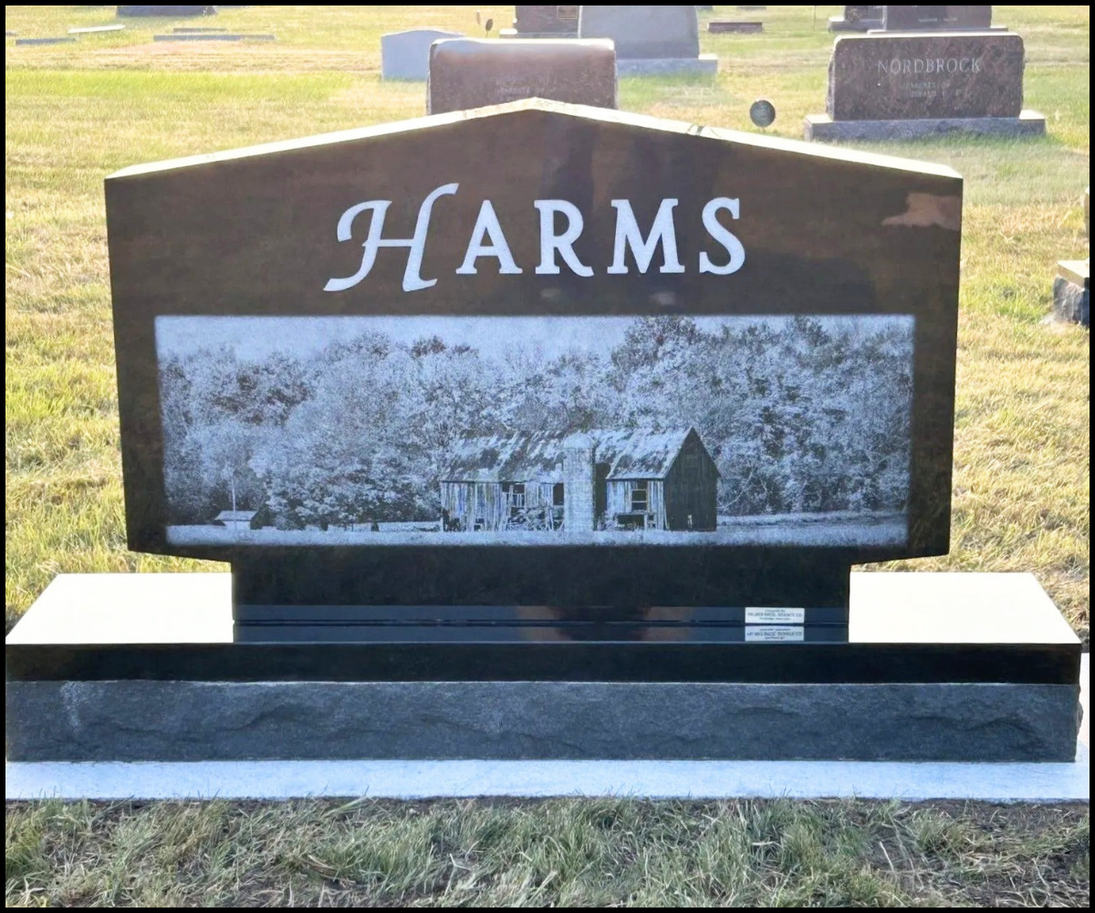 Gravestone in a cemetery with the word 'HARMS' and an engraved image of an old barn.