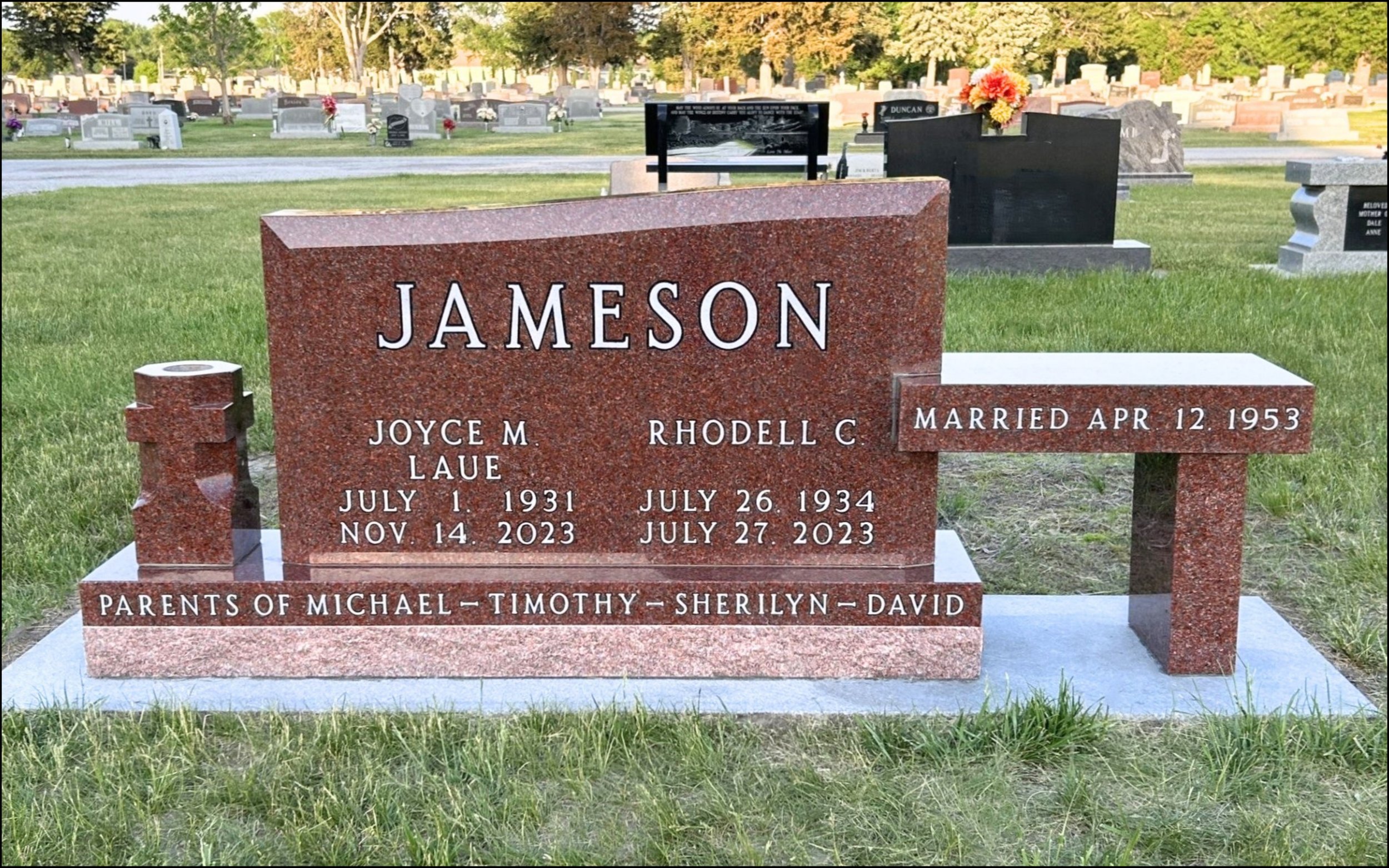 Red granite headstone in a cemetery marking the grave of Jameson, with inscription of birth and death dates, and names of his parents, Joyce M. Laue and Rhodell C. Jameson, along with space for their names and the verse about parents of Michael, Timothy, Sherilyn, and David. In the background are various other headstones and flowers in a well-kept cemetery.
