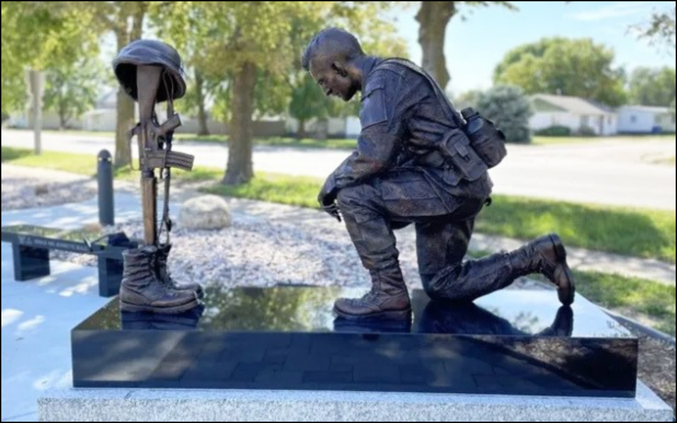 Man kneeling on the ground in a park, looking at a pair of black combat boots with rifle and helmet on the ground in front of him.