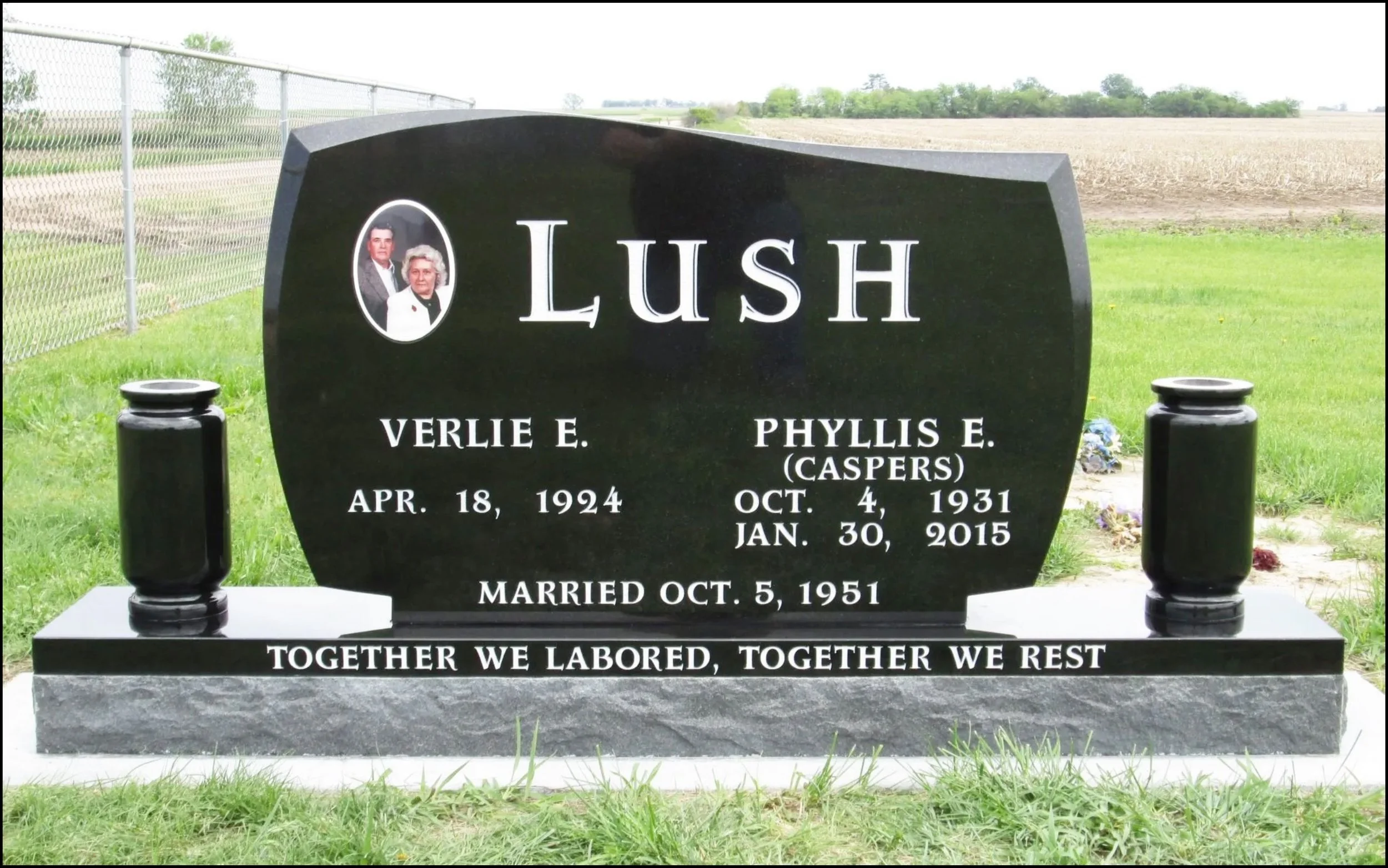 Gravestone with photos of Verlie E. and Phyllis E. (Caspers) Lush, with birth and death dates, and an inscription reading "Together we labored, together we rest." Surrounded by grass and trees in the background.