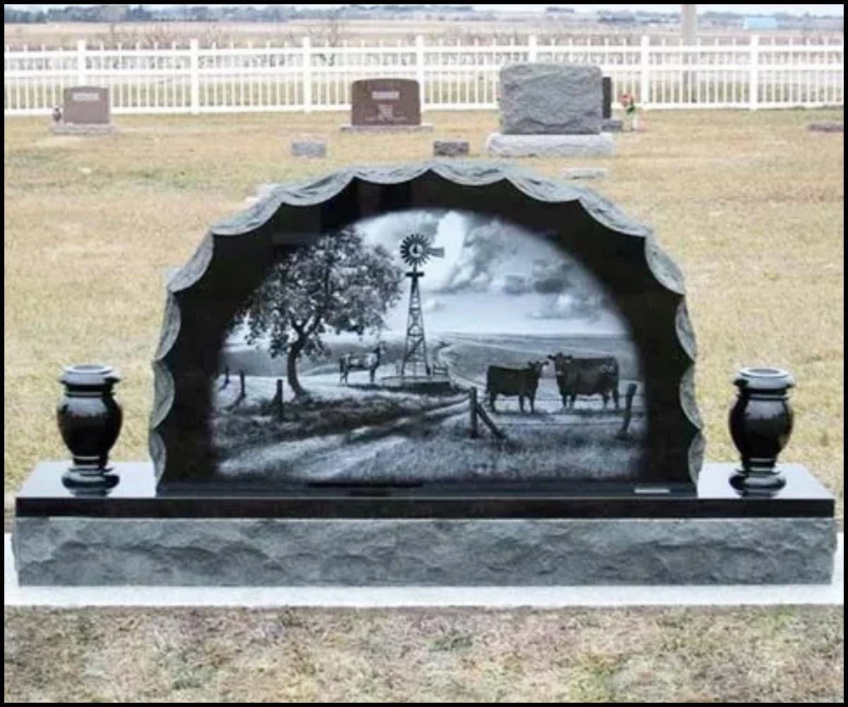 Gravestone with an etched black and white rural scene, featuring a tree, a windmill, a dirt road, and cows, set in a cemetery with a white fence in the background.
