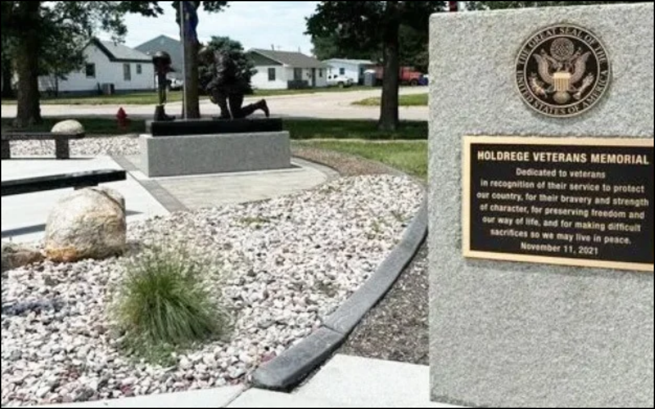 A memorial site dedicated to veterans, with a large stone plaque featuring the Great Seal of the United States ephemed on the upper right side. A black metal plaque below reads: 'HOLDREGE VETERANS MEMORIAL. Dedicated to veterans in recognition of their service to protect our country, for their bravery and strength at character, for preserving freedom and our way of life, and for making difficult sacrifices so we may live in peace. November 11, 2021.' The site includes a kneeling statue of a soldier, surrounded by landscaped rocks and small plants, with a residential neighborhood visible in the background.