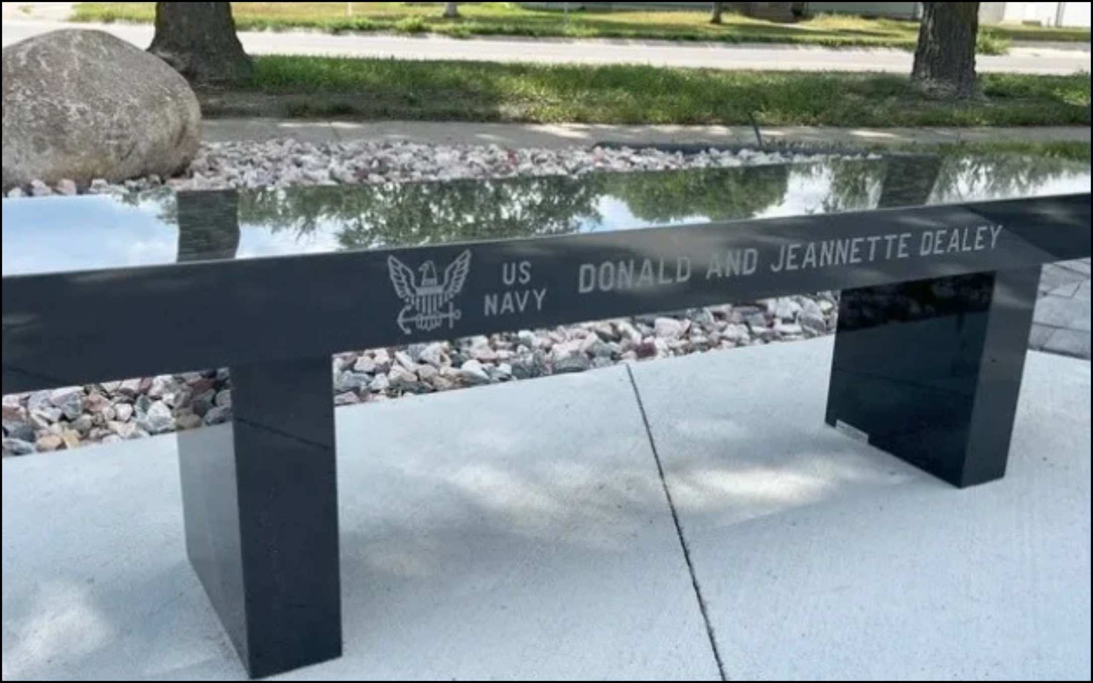 A memorial plaque dedicated to Donald and Jeannette Dealey, featuring the U.S. Navy emblem, located outdoors on a black stone bench with a reflective surface, surrounded by trees, grass, and a sidewalk.