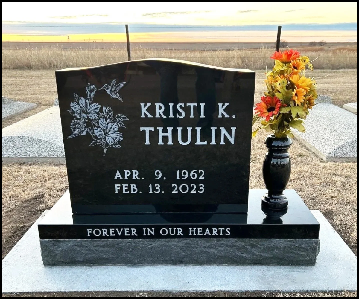 Black granite headstone with engraved white text for Kristi K. Thulin, born April 9, 1962, died February 13, 2023, with a floral illustration and a hummingbird, a black vase with yellow and orange artificial flowers on the right, and a concrete border with the inscription "Forever in Our Hearts" at a cemetery.