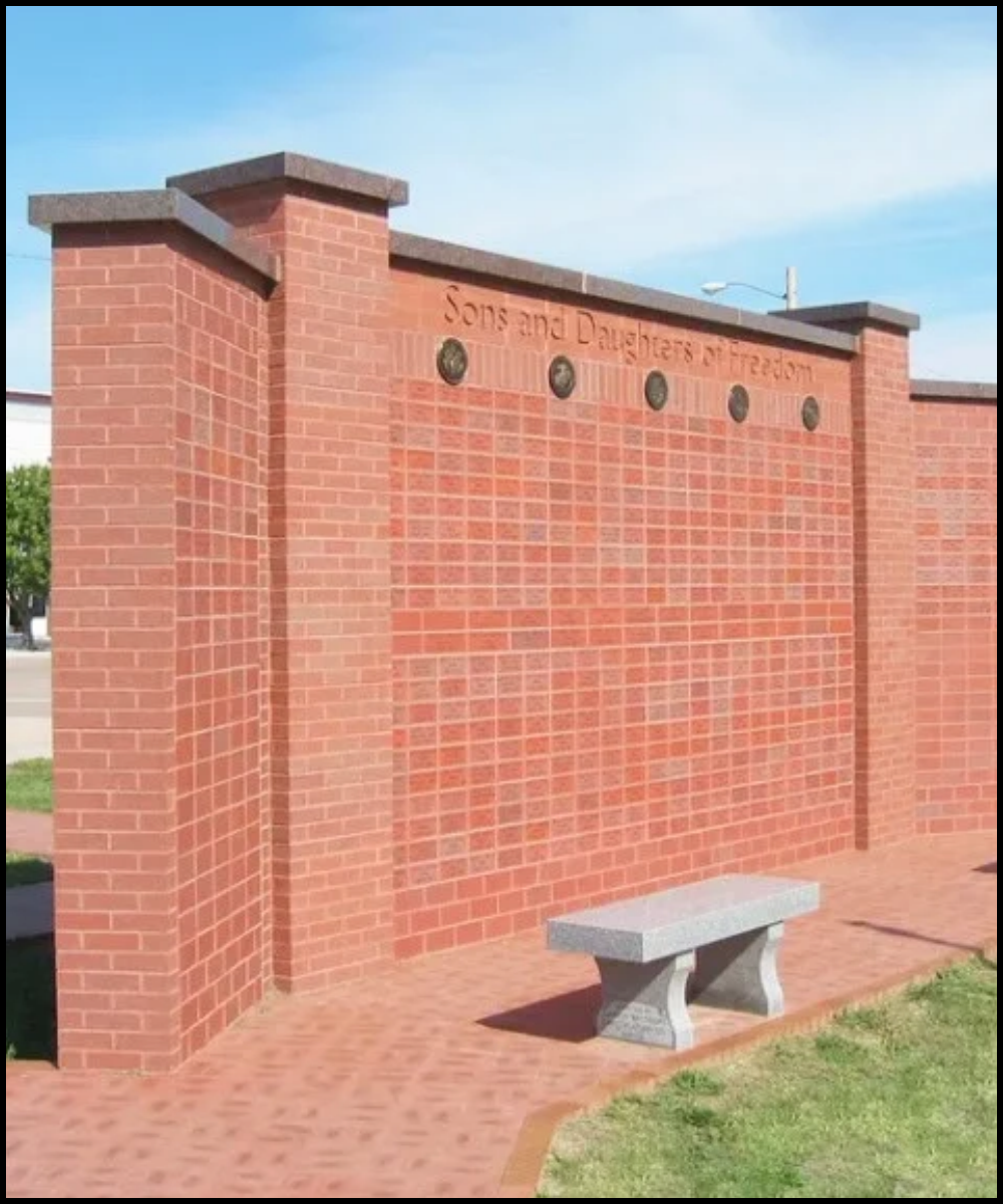 A brick memorial wall with an inscription 'Sons and Daughters of Freedom' at the top, featuring several plaques and a stone bench in front.