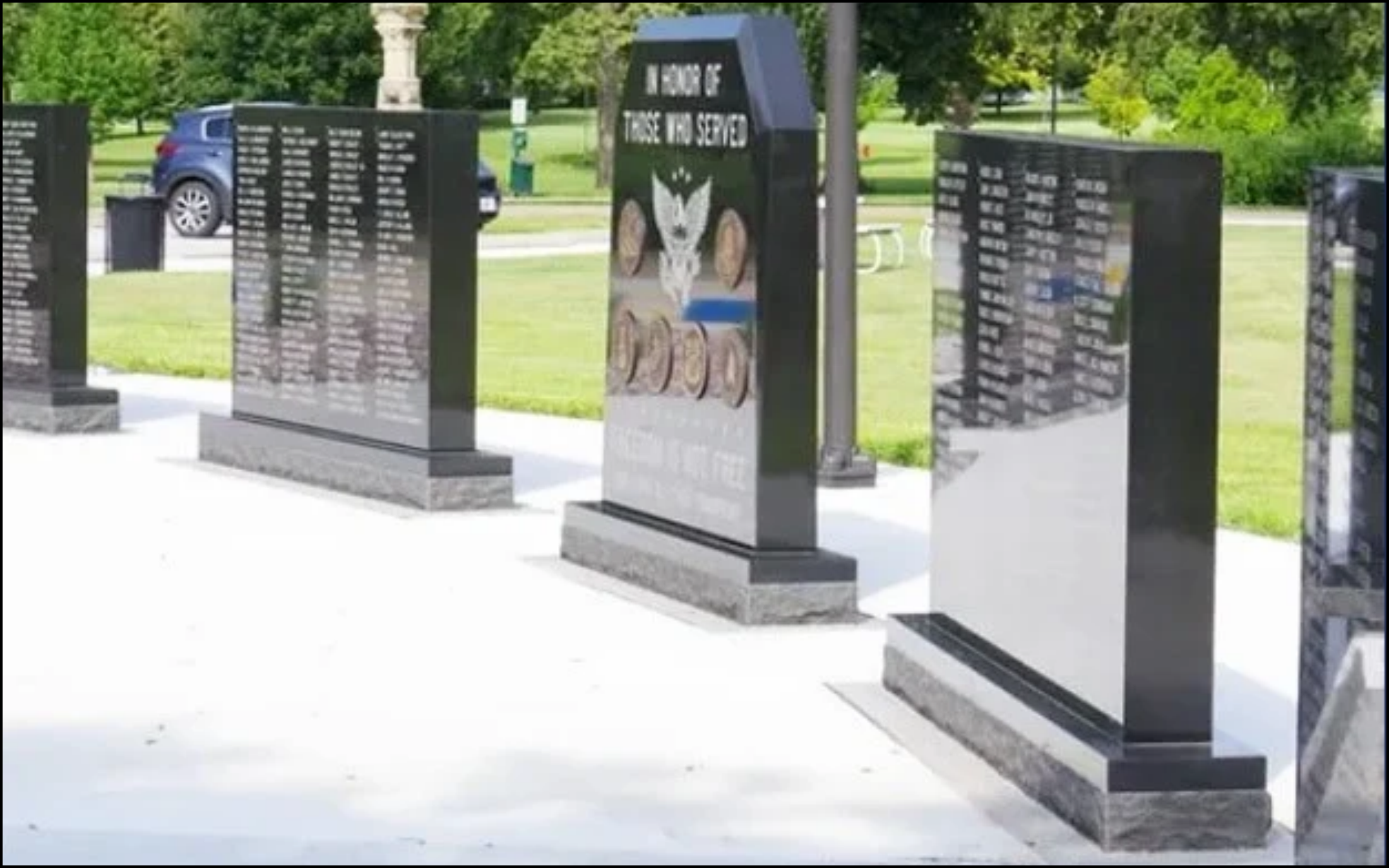 A memorial wall with multiple black granite plaques inscribed with names, situated in a park with green grass and trees. The wall has a central panel with the words 'IN HONOR OF THOSE WHO SERVED' and an eagle emblem.