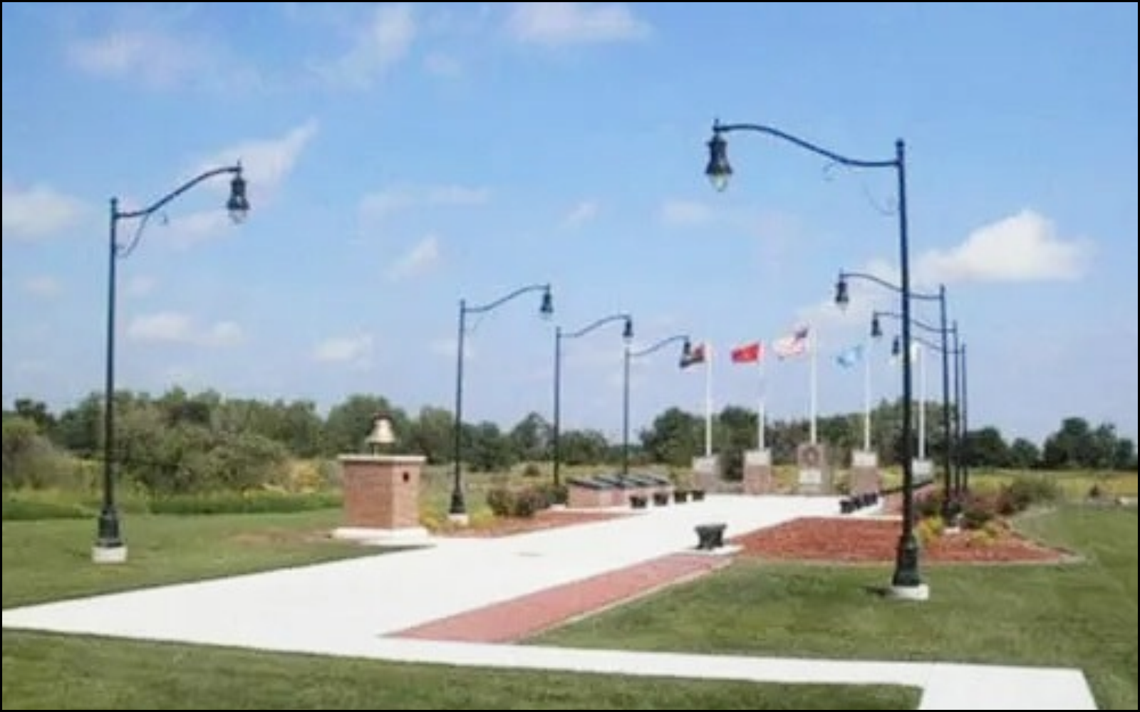 A park with pathway, street lamps, flags, benches, and a green grassy area.