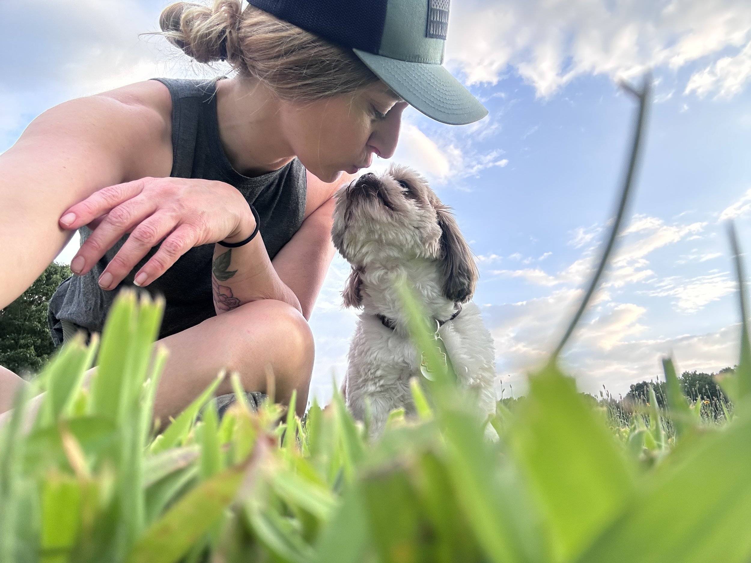 A person with a baseball cap and tank top is close to the ground, holding a small dog, in a grassy outdoor setting under a partly cloudy sky.