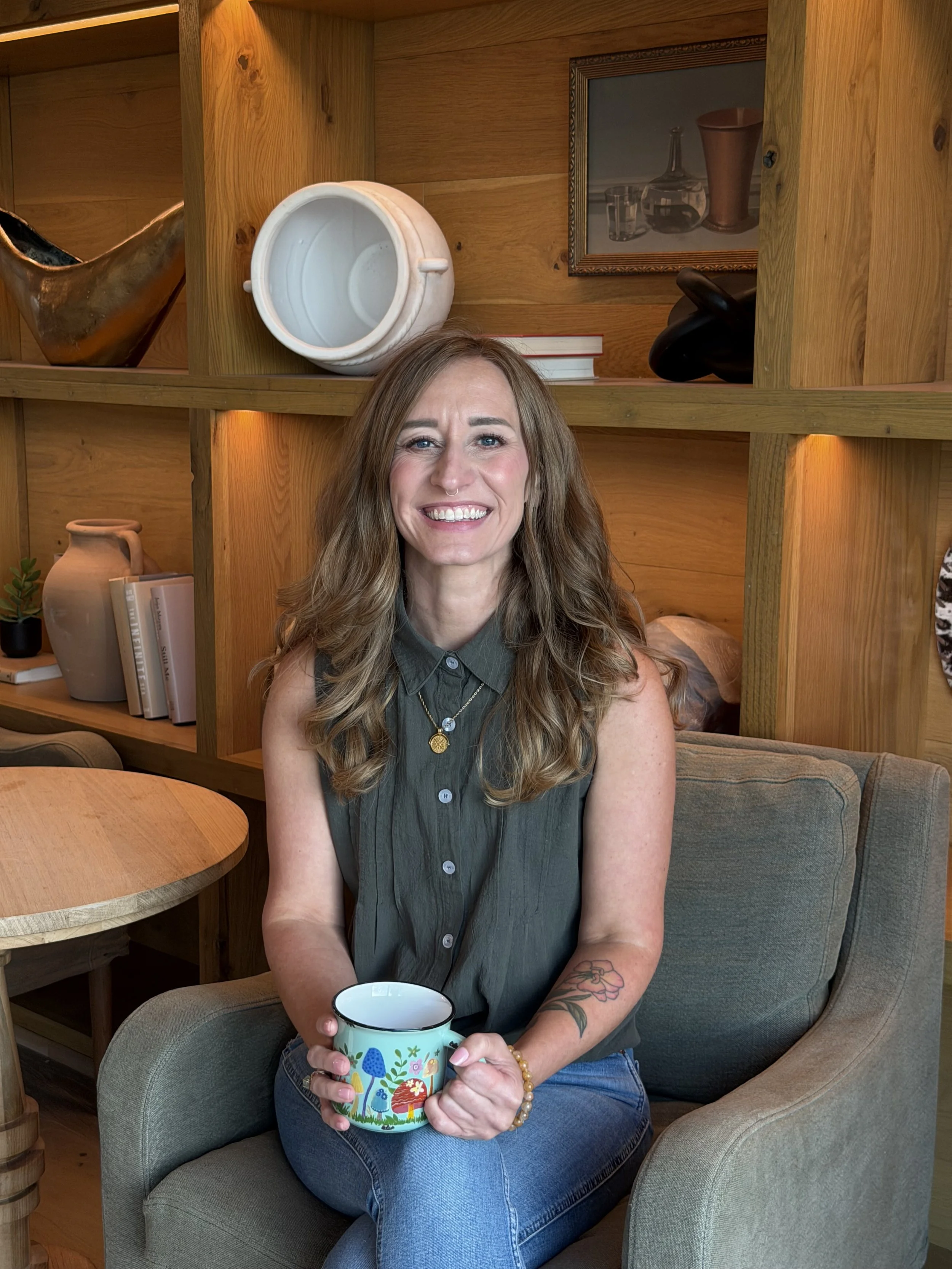 A woman with long wavy hair, smiling, sitting in a green armchair, holding a colorful mug with a floral design, in a cozy wooden interior.