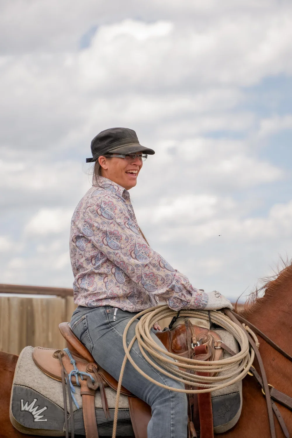 Smiling woman wearing a cowboy hat and riding gear sits on a saddled horse with a coiled lasso.