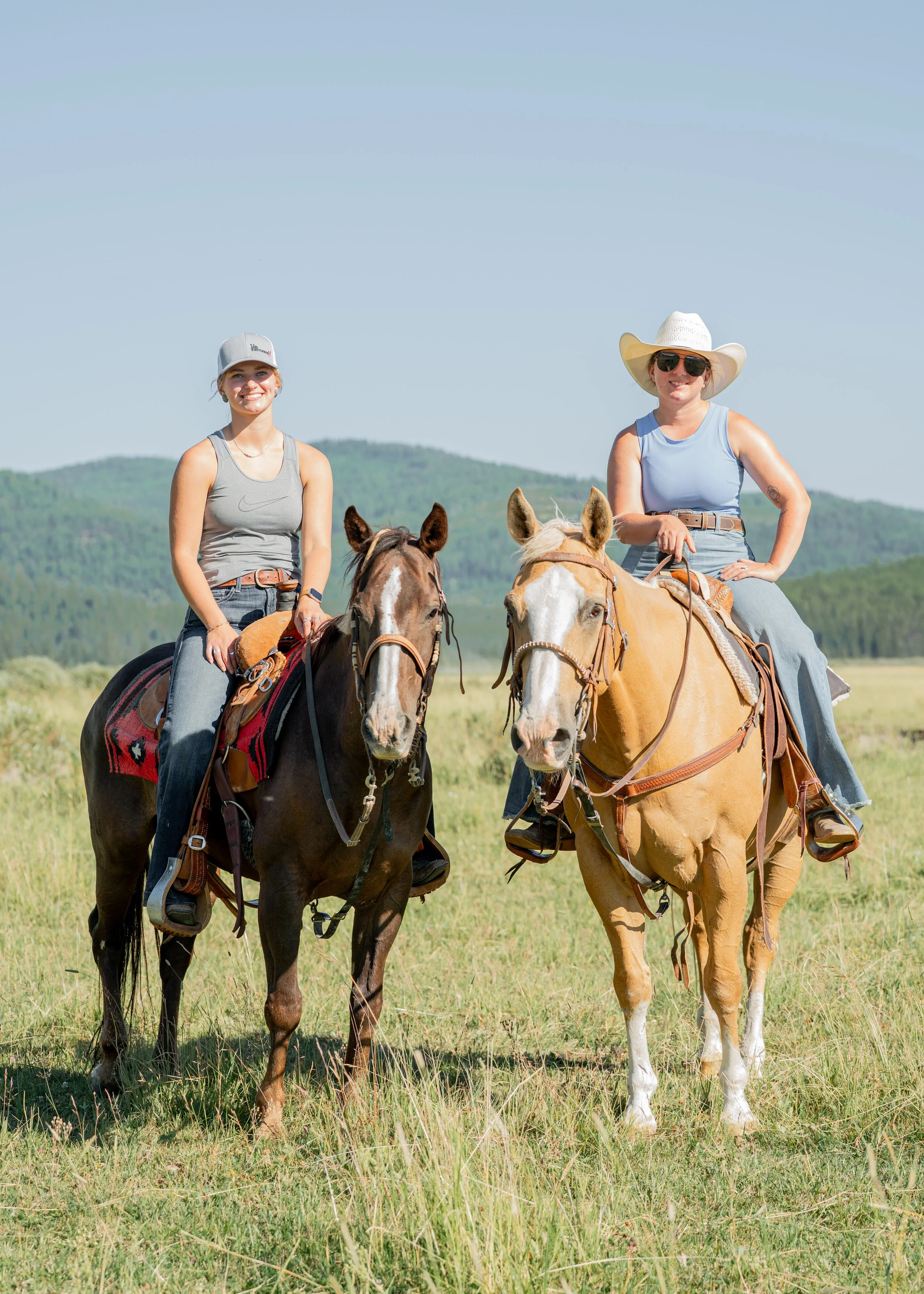 Two women riding horses through a grassy mountain meadow under a clear blue sky