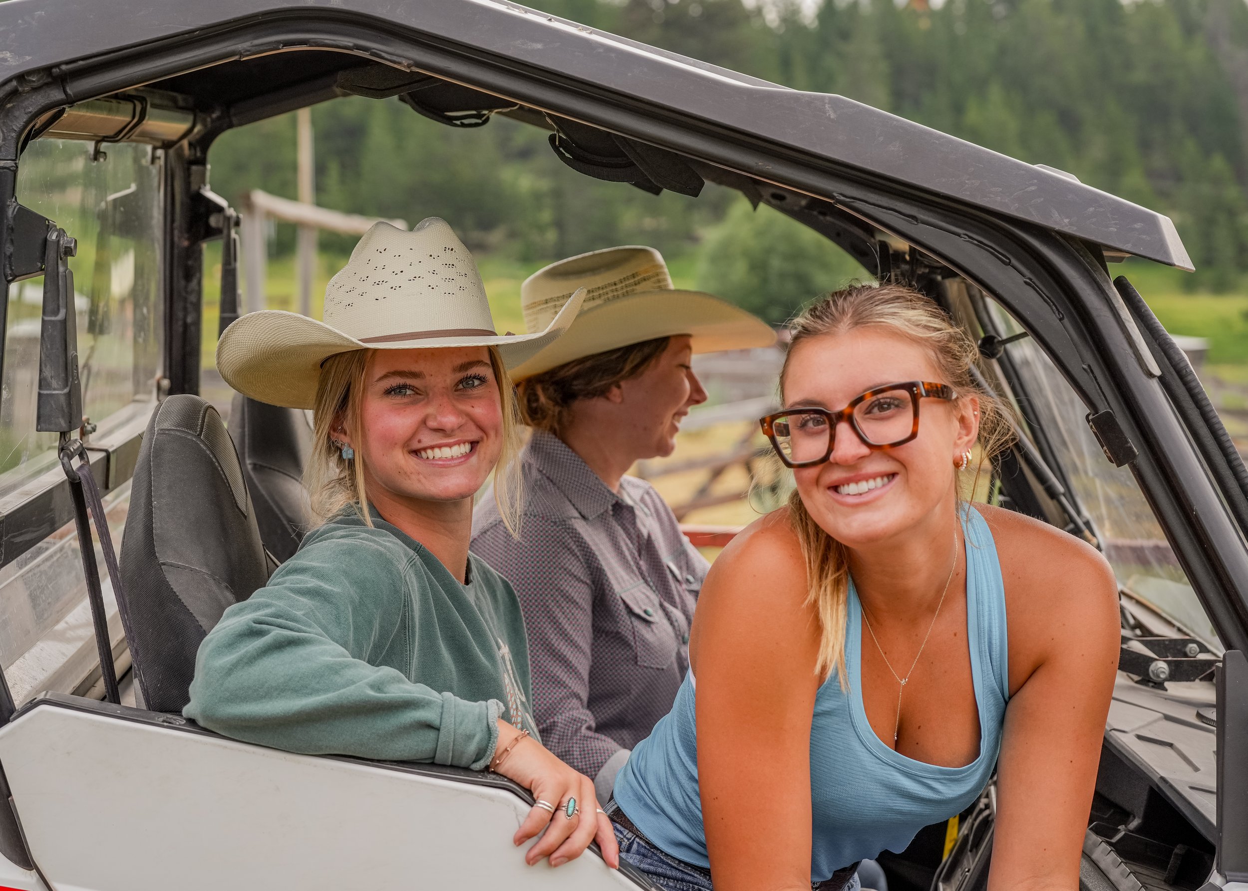 Three friends smiling for a selfie in an open-sided off-road vehicle outdoors, with two wearing cowboy hats.
