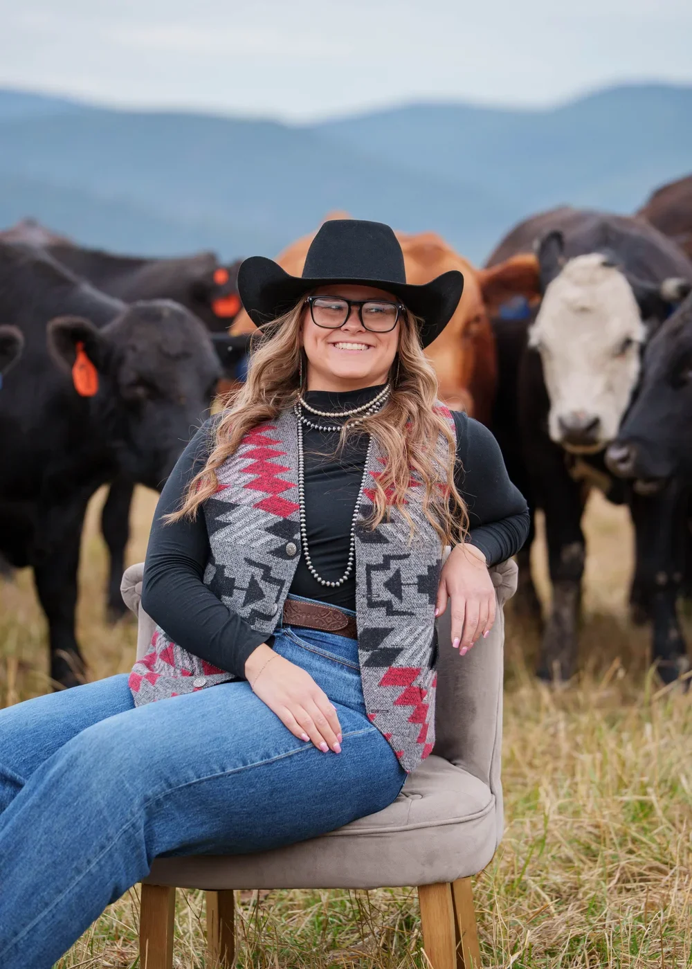 Smiling woman in a black cowboy hat, glasses, and patterned vest seated in front of a herd of cattle