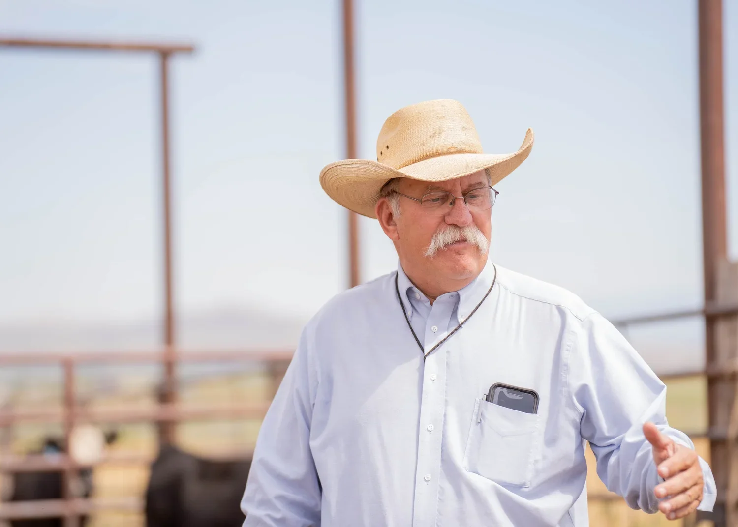 Older man wearing a cowboy hat and glasses standing outdoors near a ranch fence