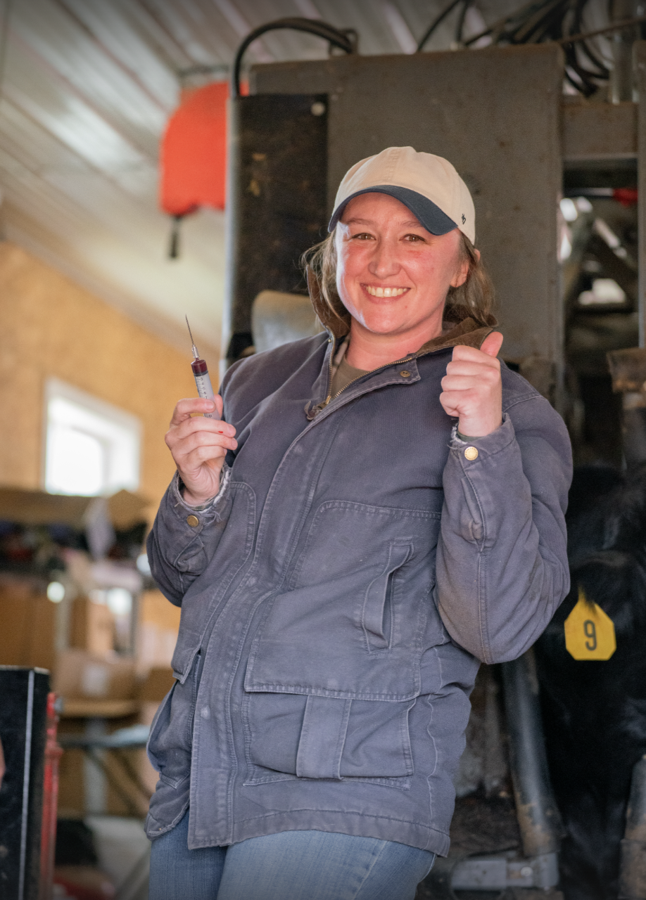 Smiling woman in a work jacket and baseball cap holding a syringe and giving a thumbs-up while working to vaccinate cattle
