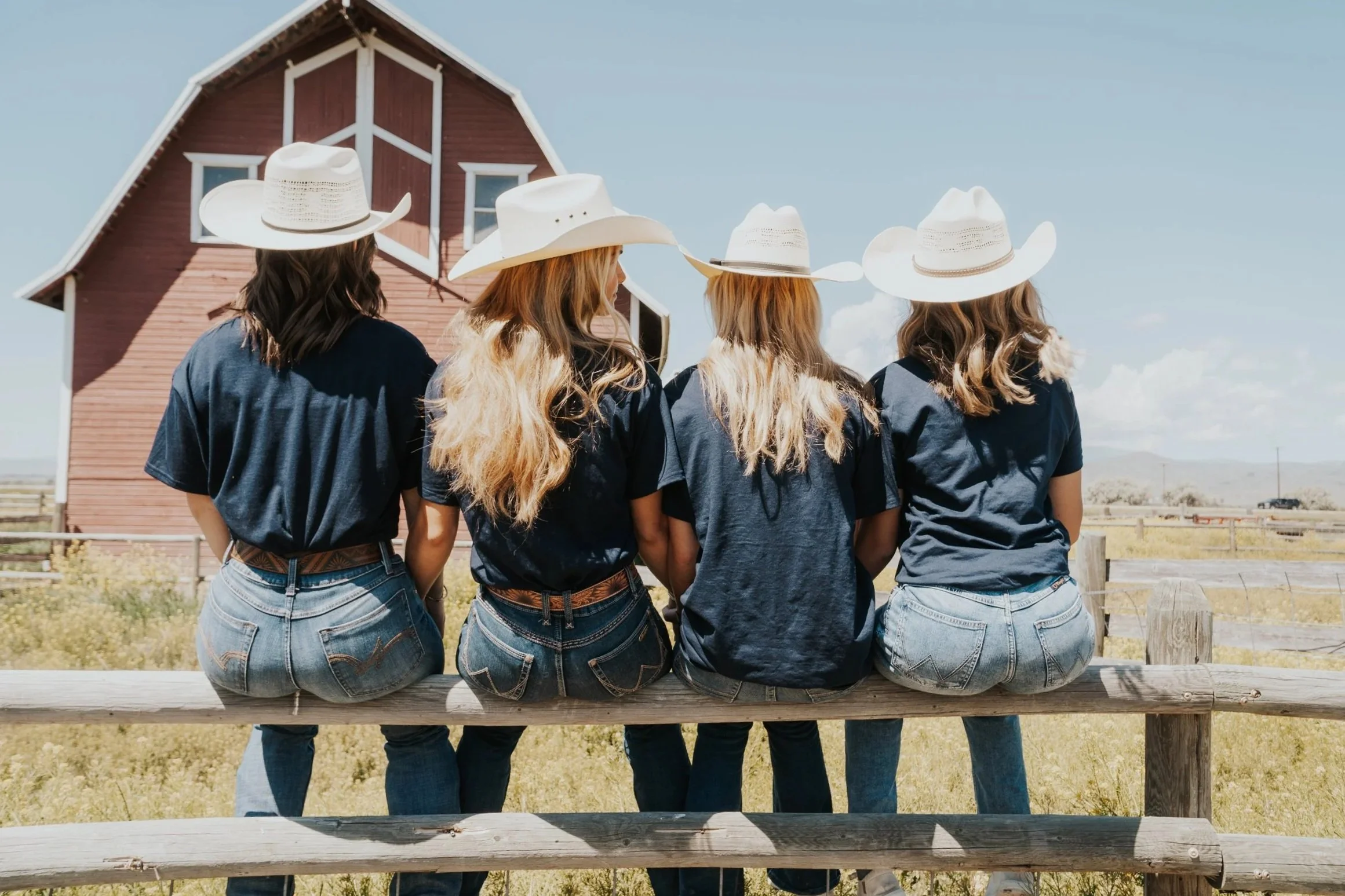 Four women in cowboy hats and jeans sitting on a wooden fence in front of a red barn