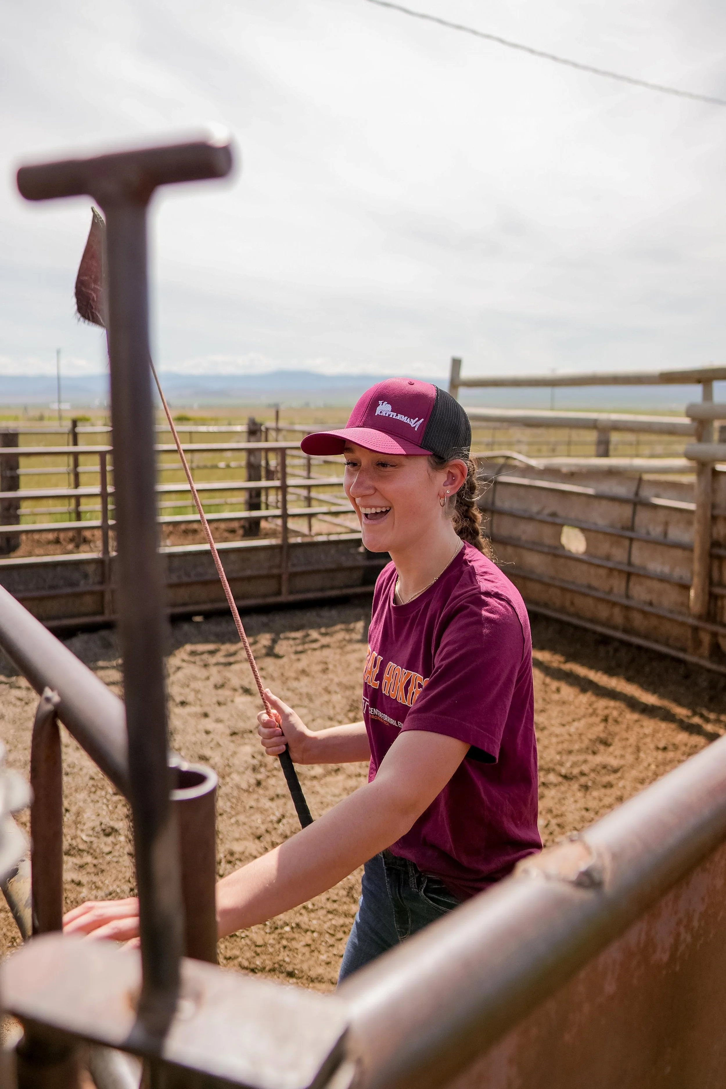 Smiling woman in a cattle ranch pen holding a rope, wearing a cattleman's cap and maroon shirt.