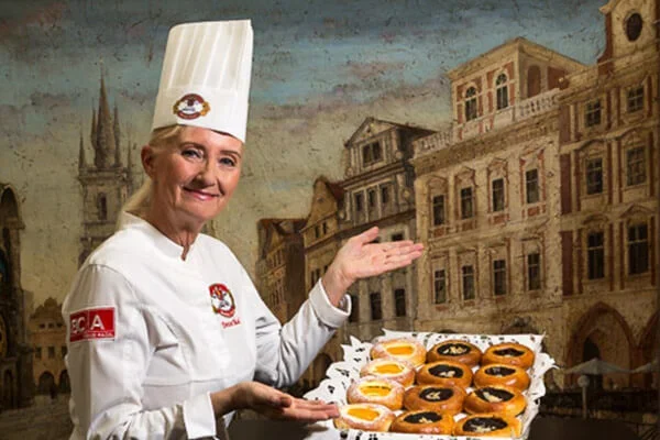 A female pastry chef in a white chef's outfit and tall hat holding a tray of assorted donuts, standing in front of a painted cityscape backdrop.