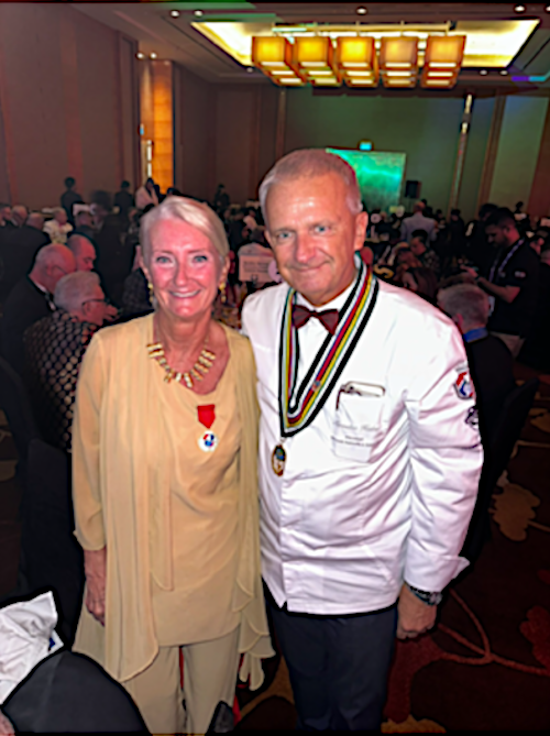 An elderly woman and a man in a chef uniform at an award ceremony, both wearing medals, in a large, well-lit banquet hall with many people seated at tables.