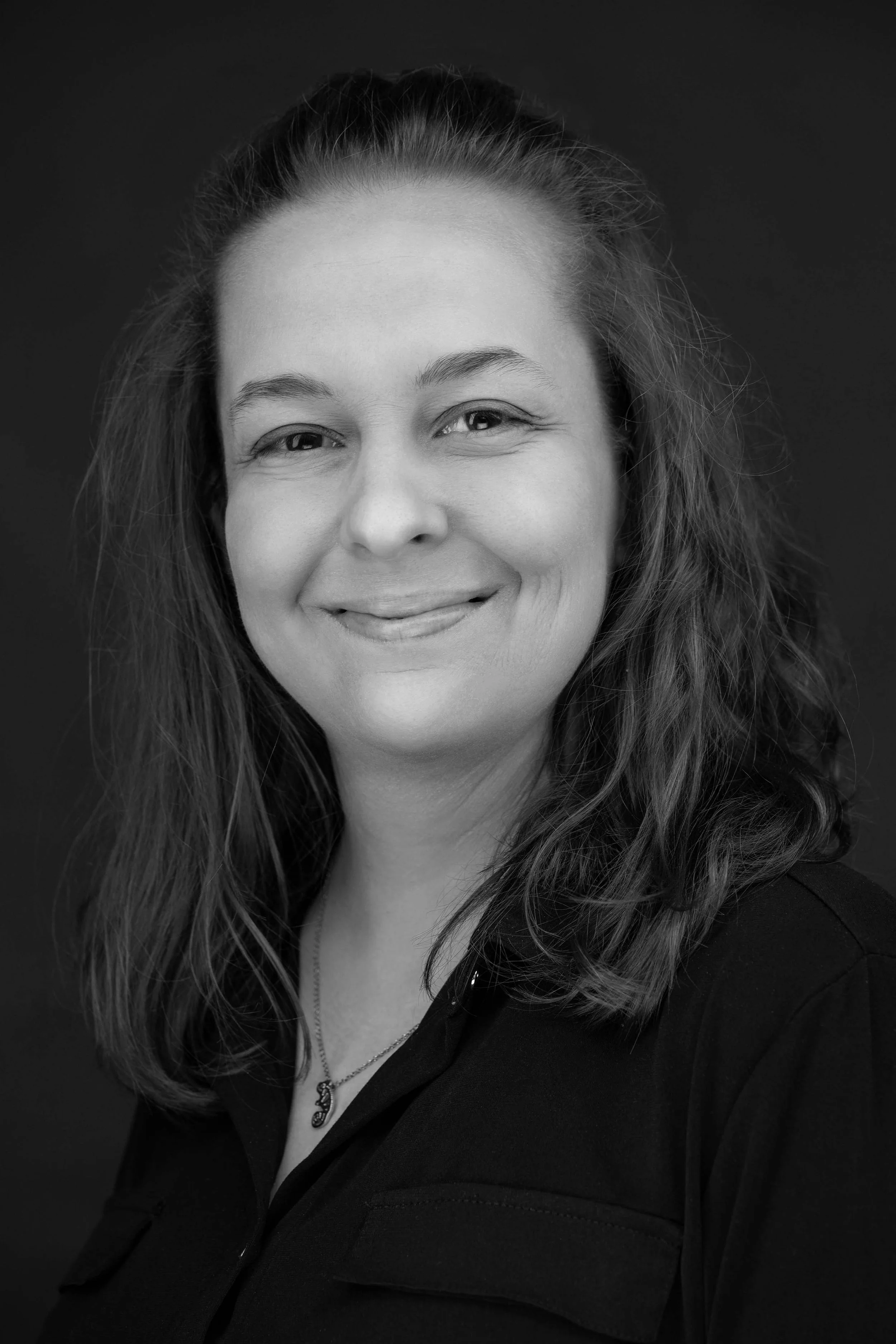 Black and white portrait of a woman with long wavy hair, smiling, wearing a necklace with a pendant, and a dark top.
