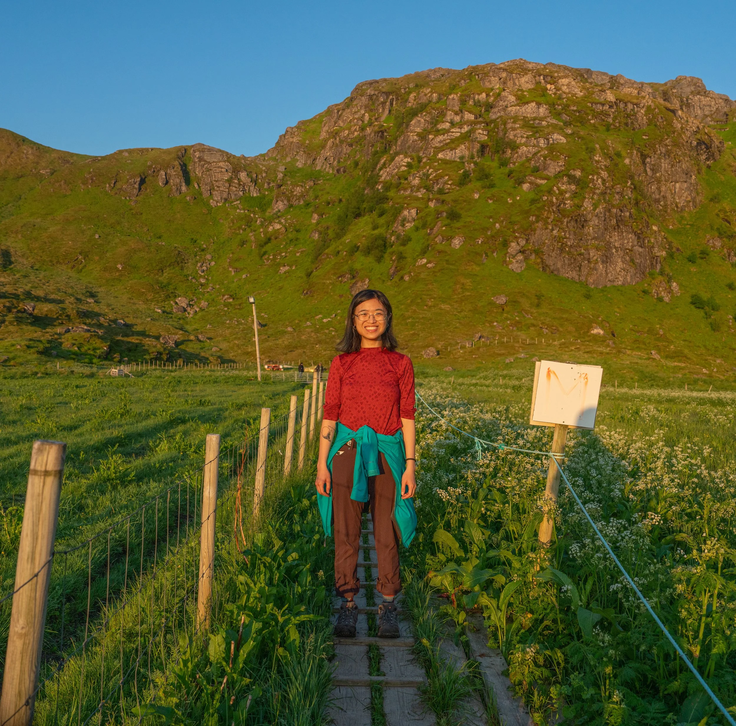 A woman smiling, wearing glasses, a red patterned top, brown pants, and hiking boots, standing on a wooden pathway surrounded by green plants and flowers, with a hill and blue sky in the background.