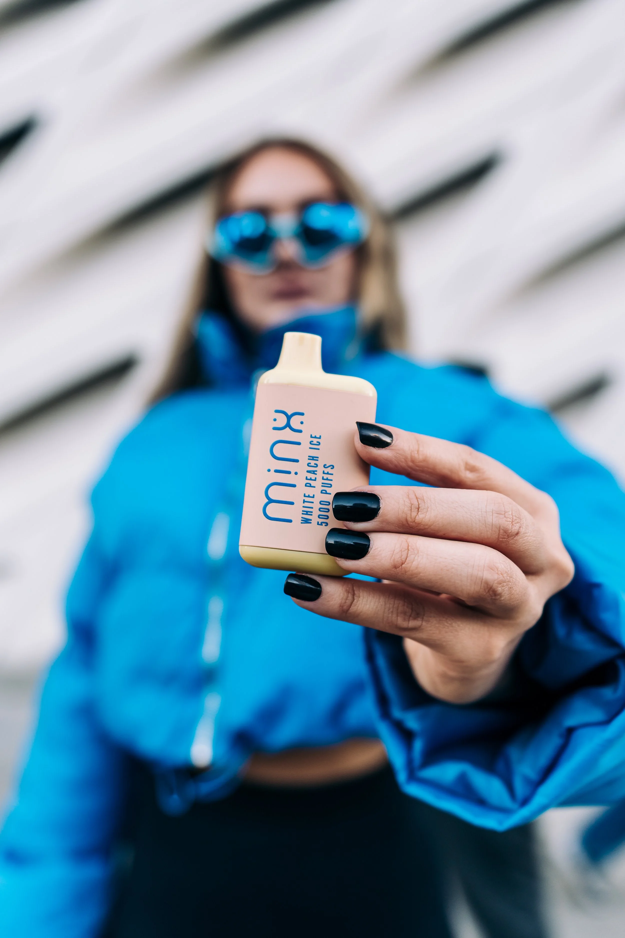 A woman in blue jacket and sunglasses holding a BUX white peach ice pop close to the camera.