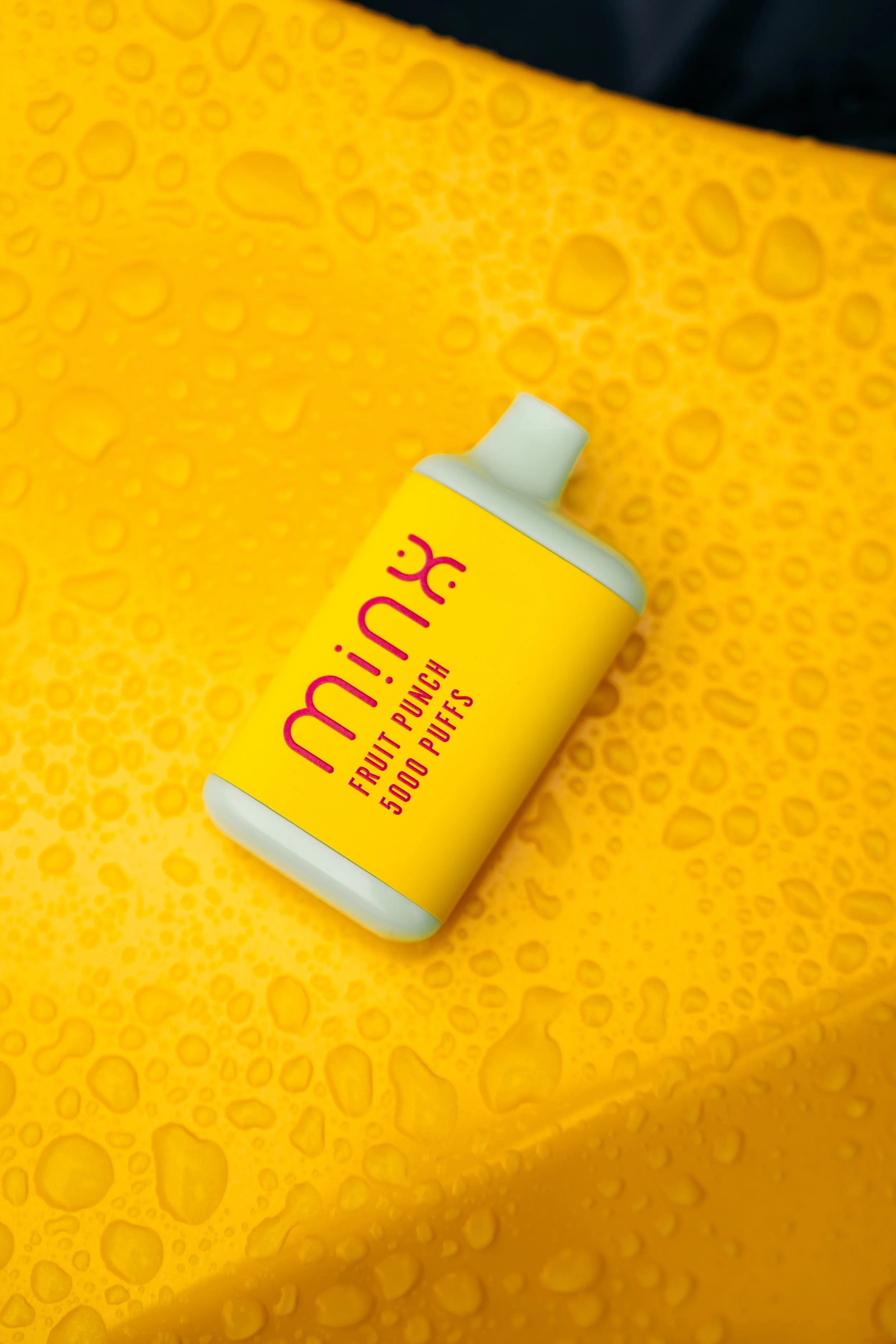 A yellow container of Mine fruit punch flavored puffs with 5000 puffs written on it, placed on a yellow surface with water droplets.