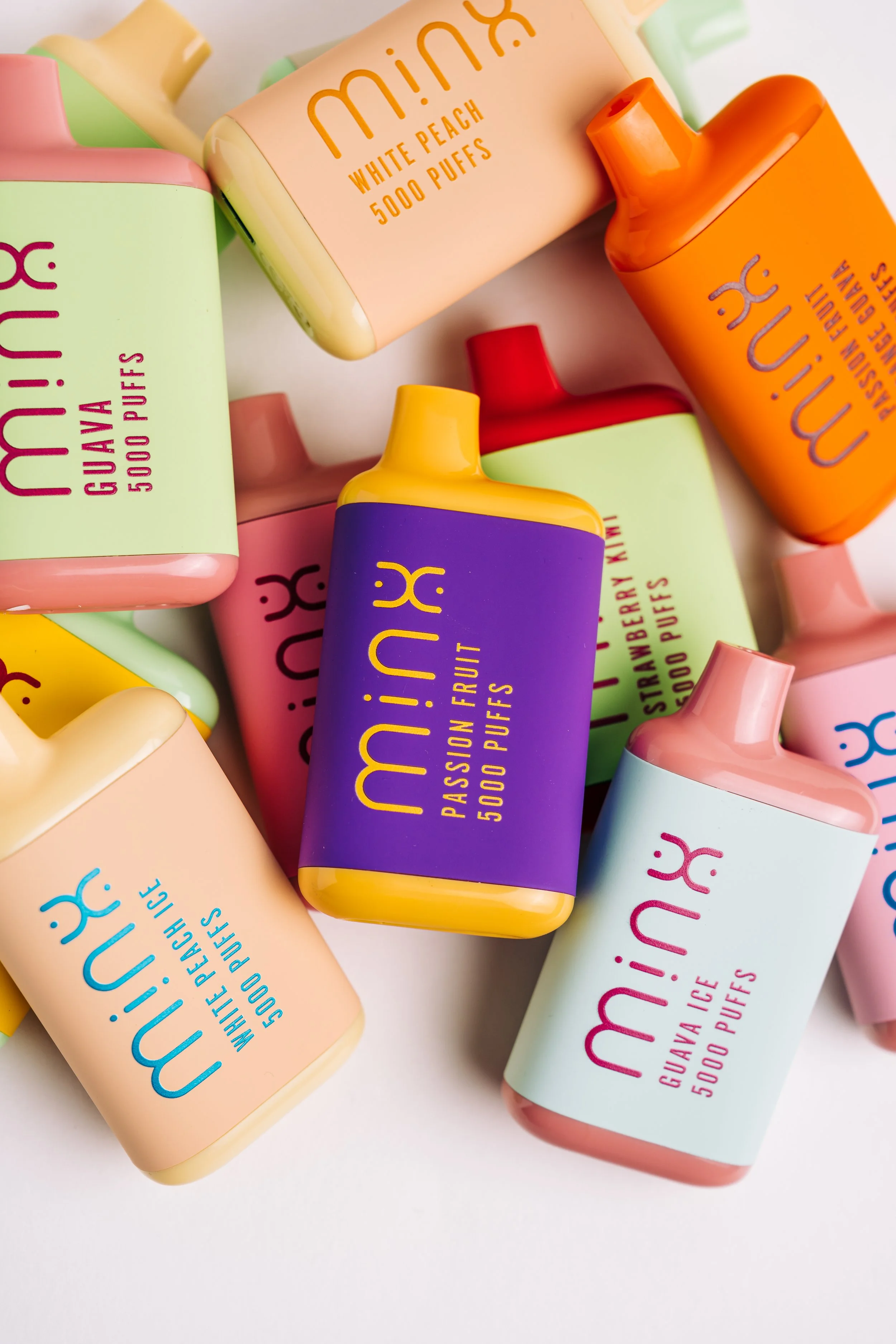 Colorful collection of small, rectangular bottles with rounded edges, labeled with fruity flavors such as white peach, passion fruit, guava, and strawberry kiwi, and a note indicating each contains 5000 puffs.