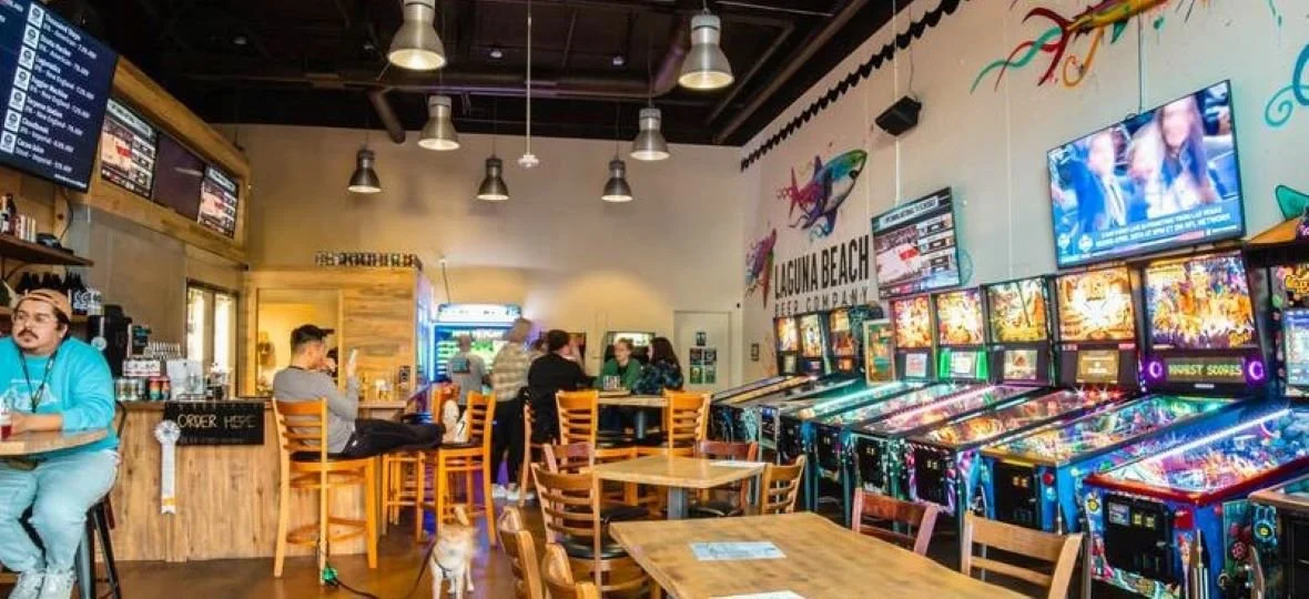 Interior of a lively arcade with pinball machines along the wall, a small bar area to the left, and people sitting on chairs and at tables, including a man with long hair and a beard. Bright wall decor features fish and the text 'Laguna Beach.'