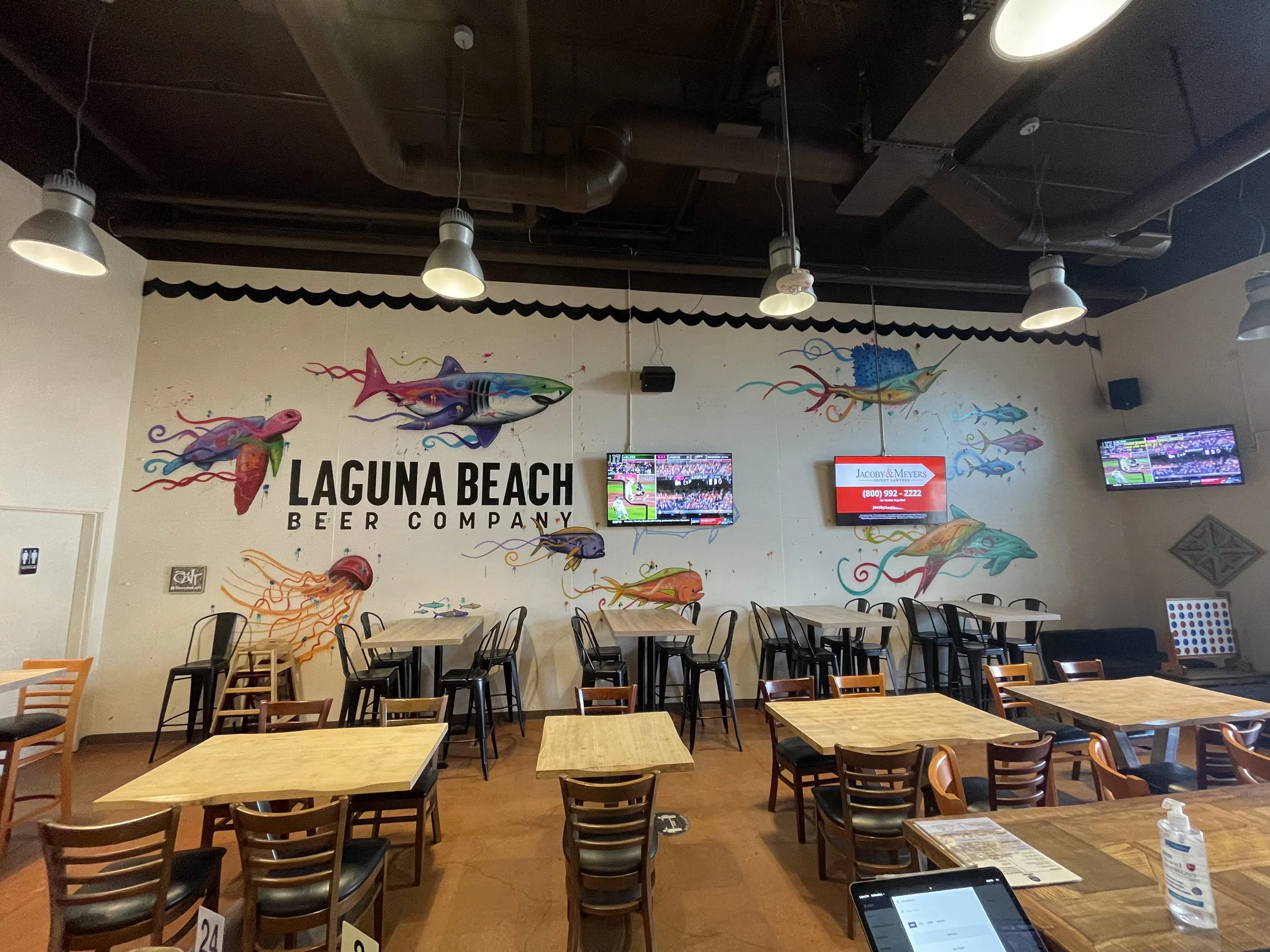 Interior of a restaurant at Laguna Beach Beer Company featuring colorful murals of fish and marine life on the wall. There are wooden tables and chairs, and multiple television screens on the wall.