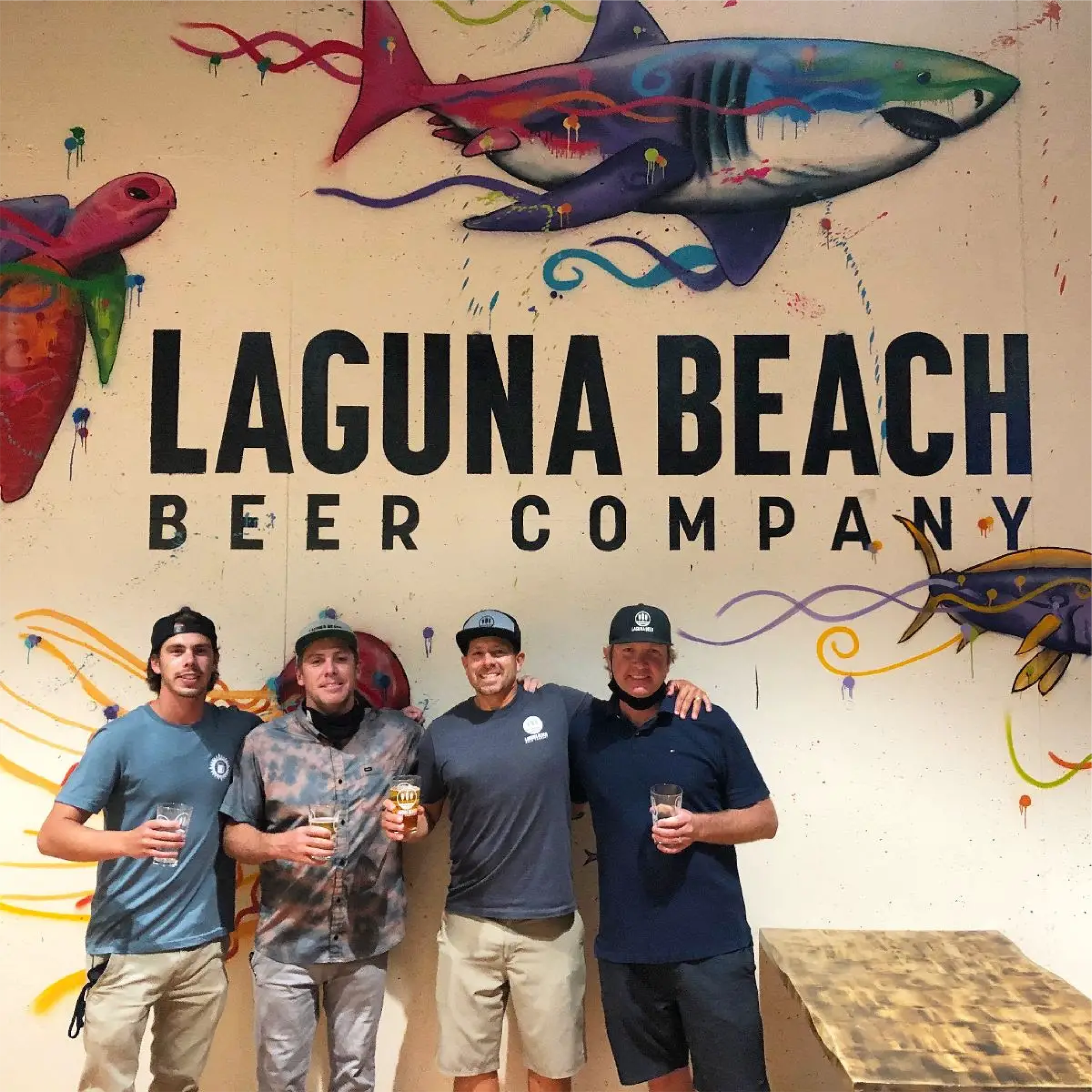 Four men standing together in front of a wall with a colorful fish mural and the text ‘LAGUNA BEACH BEER COMPANY,’ holding glasses of beer and smiling.