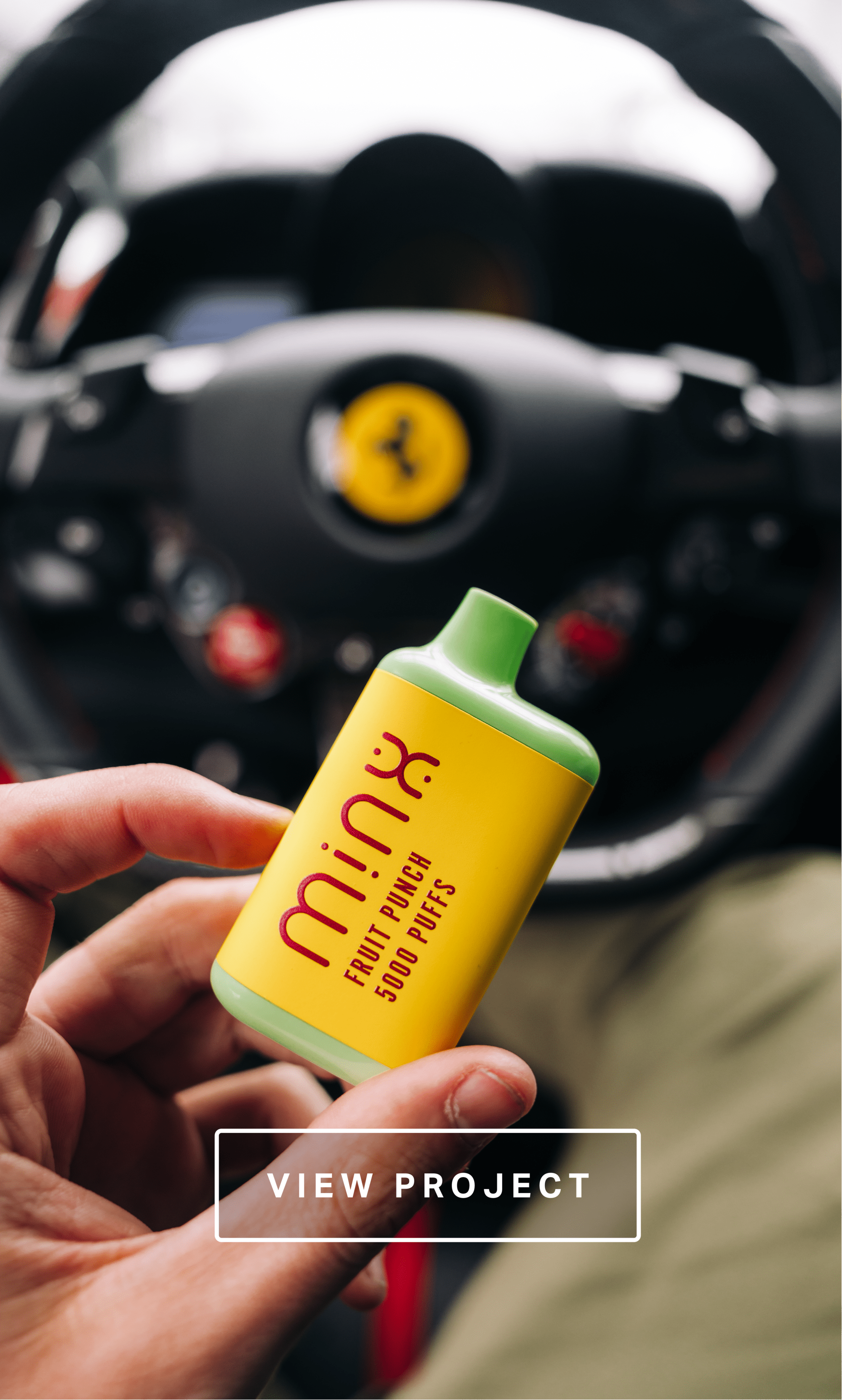 A hand holding a yellow and green pack of fruit punch flavored Puff bars in front of a blurry black steering wheel with a yellow Ferrari logo.