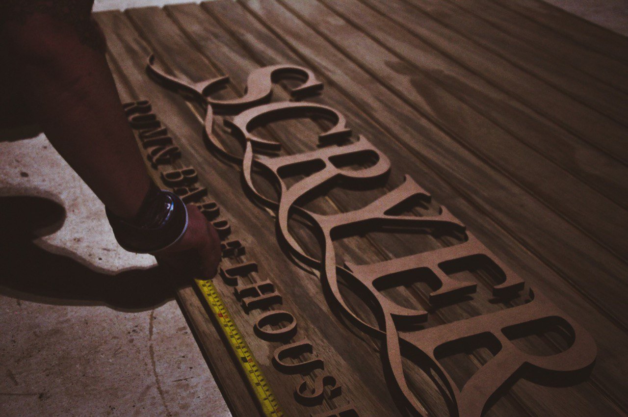 Person measuring and installing wooden sign with black cursive lettering on wood surface.