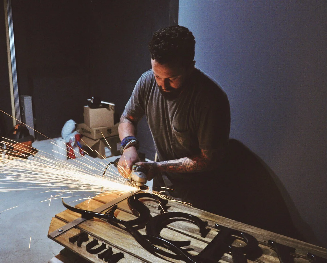 A man with tattoos working with a grinding tool, sparks flying, on a wooden sign that reads 'TRUMP' mounted on a wooden table.