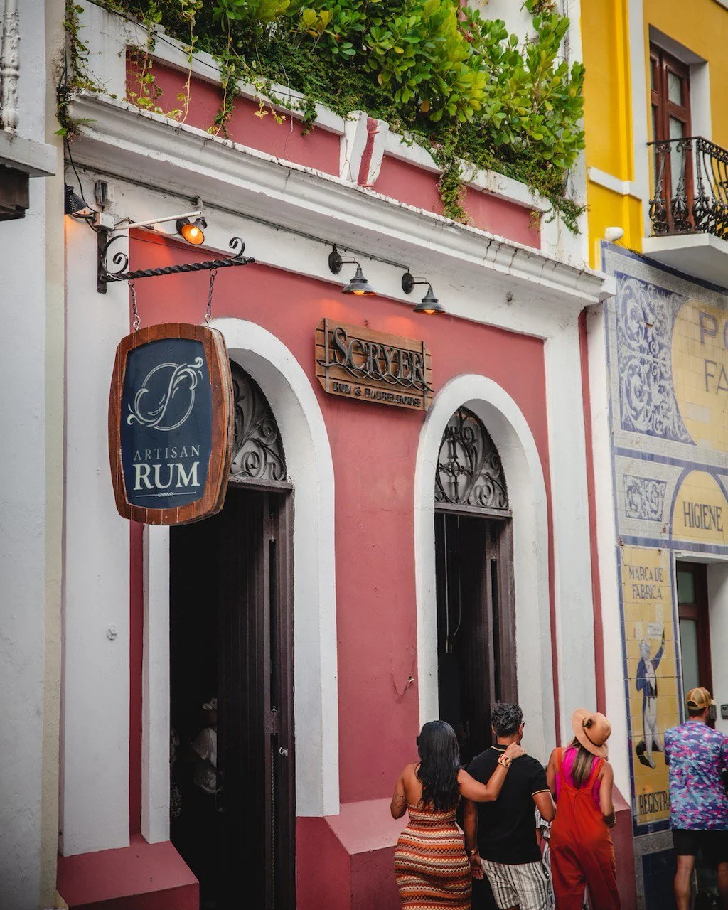 People walking past a colorful building with a sign that reads 'ARTISAN RUM' and another sign above the doorway that reads 'SERVER NOW & BARRELHOUSE' in a lively city street.