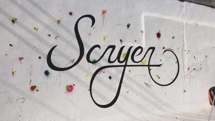 A mural with the word 'Suyen' written in black script on a white wall, decorated with colorful paint splatters and drips.